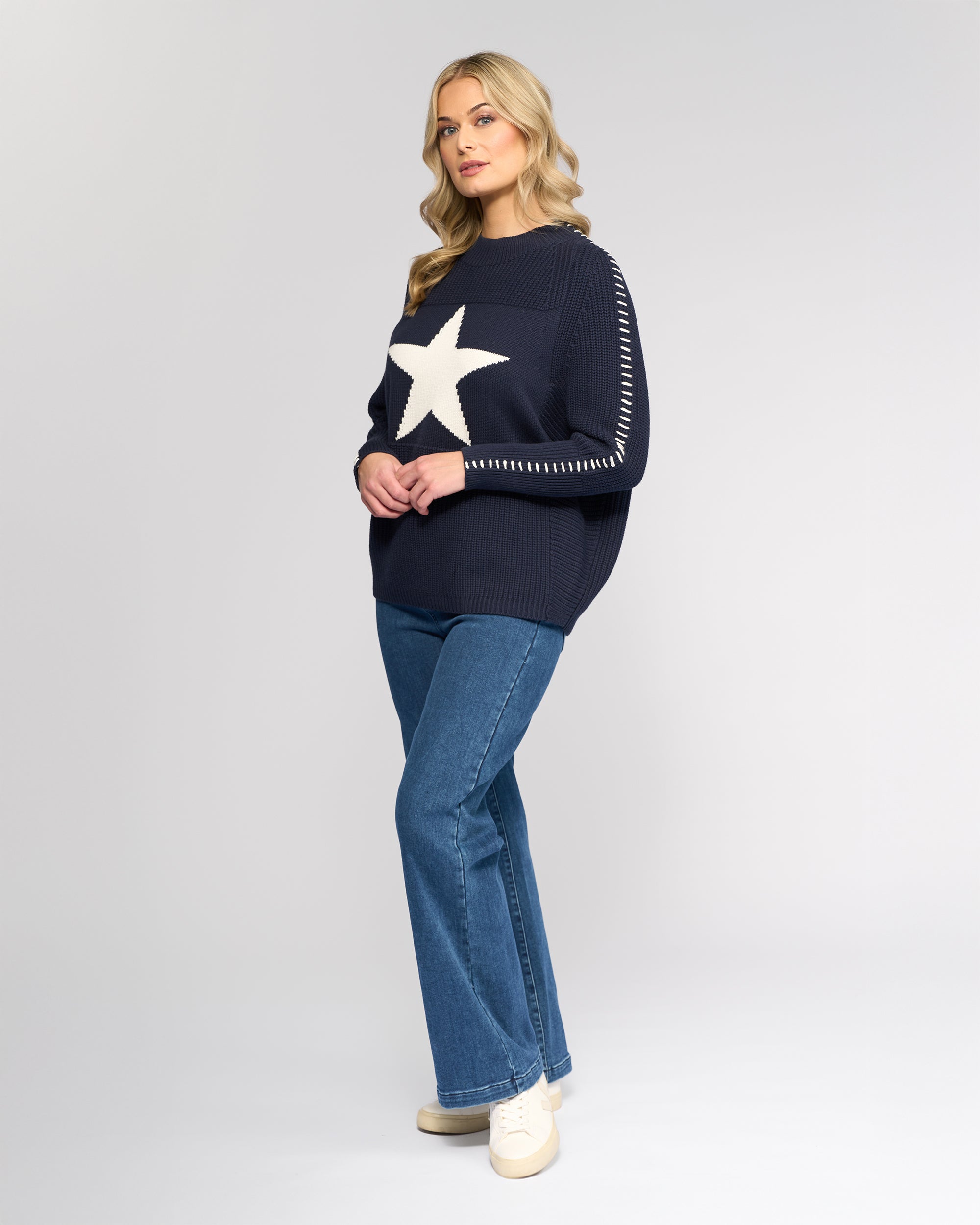 A woman with long blonde hair stands against a plain background wearing the Vassalli Star Sweater - Ink, featuring a large white star on the front, paired with blue jeans and beige sneakers. She is looking slightly to the side.