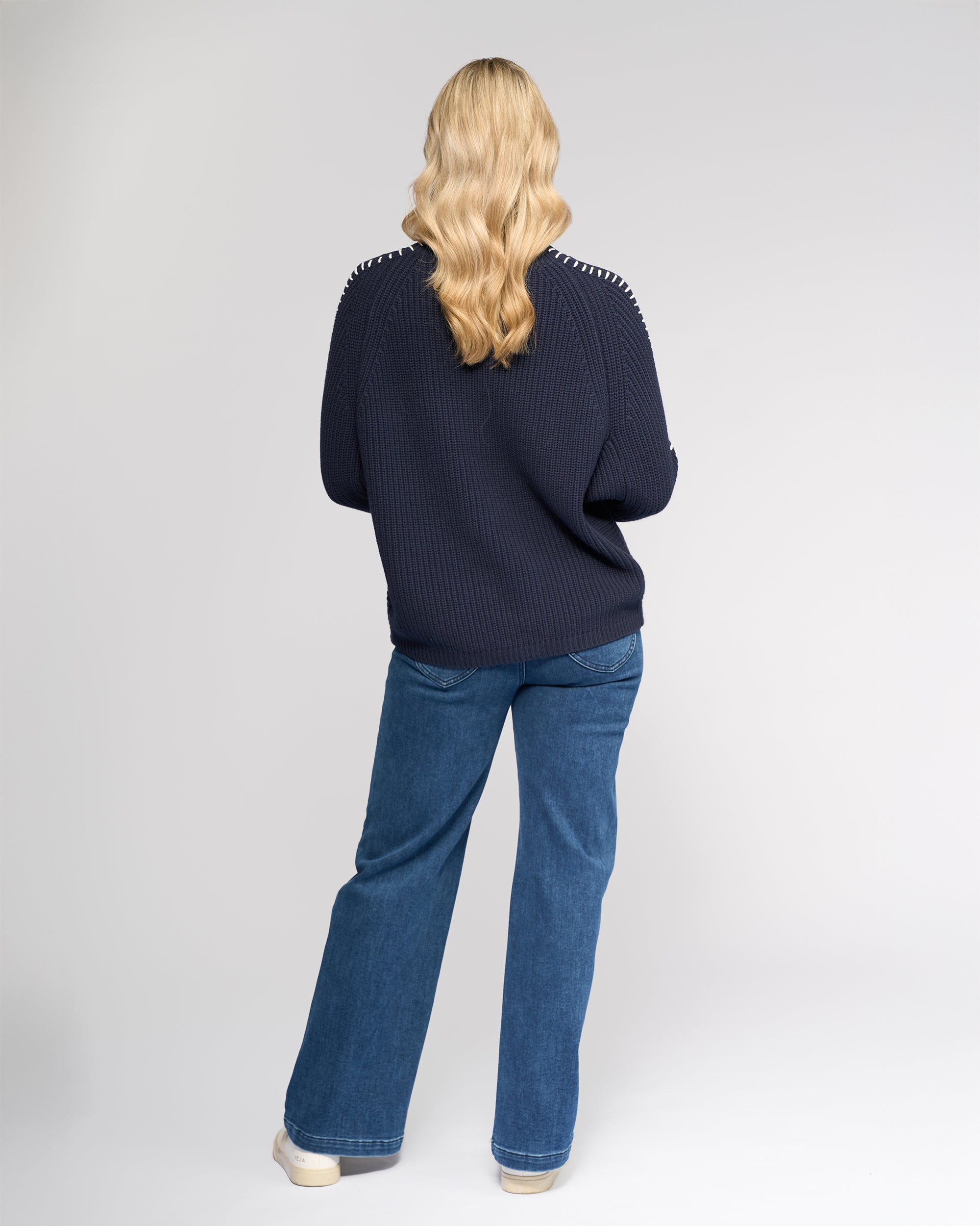 A person with long wavy blonde hair, wearing the Vassalli Star Sweater - Ink and blue jeans, stands facing away from the camera against a plain light background.