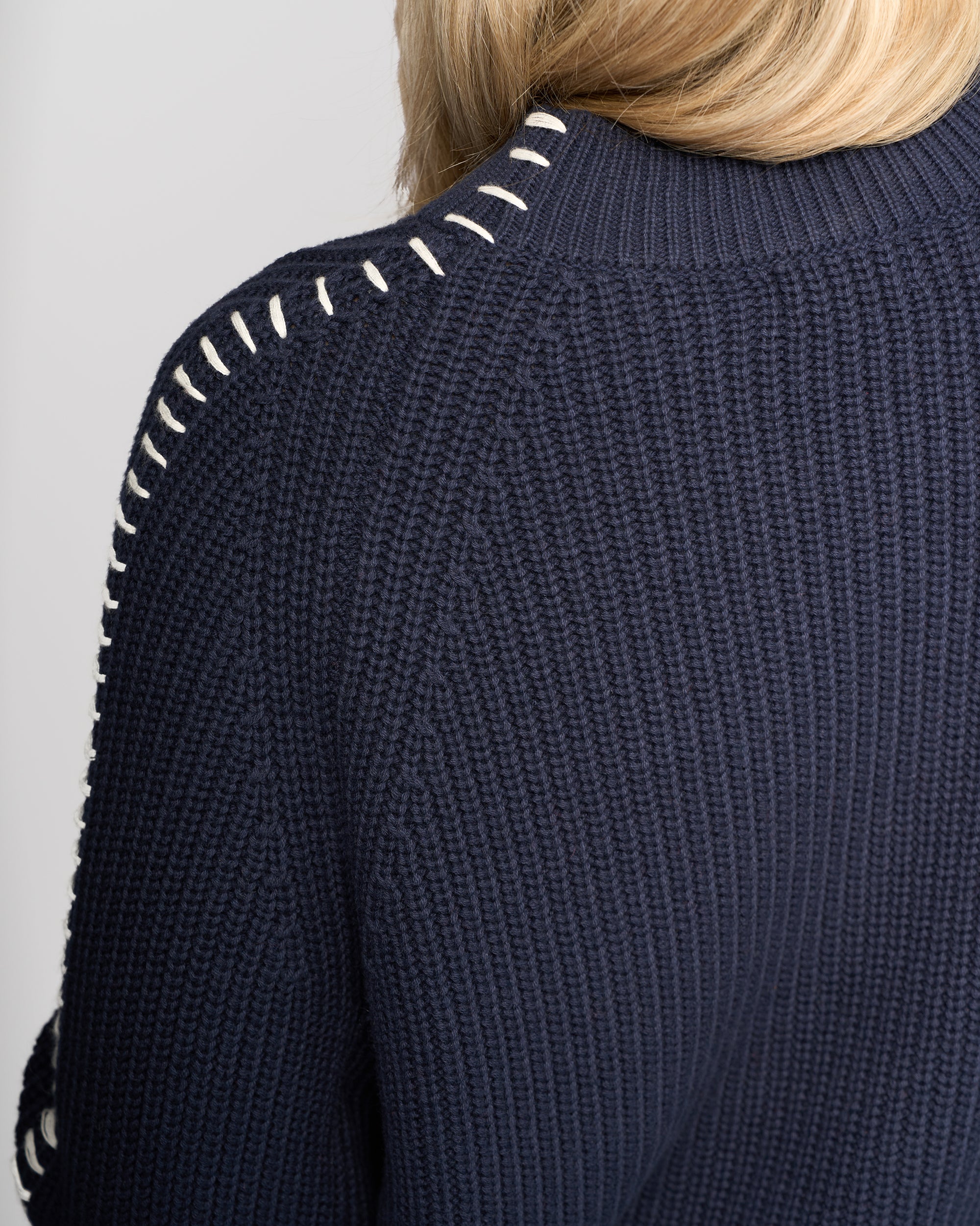 Close-up of a person wearing the Vassalli Star Sweater - Ink, an oversized rib-knit design in navy blue with white whipstitch detailing on the shoulder seam. Only the back and part of their blonde hair are visible.