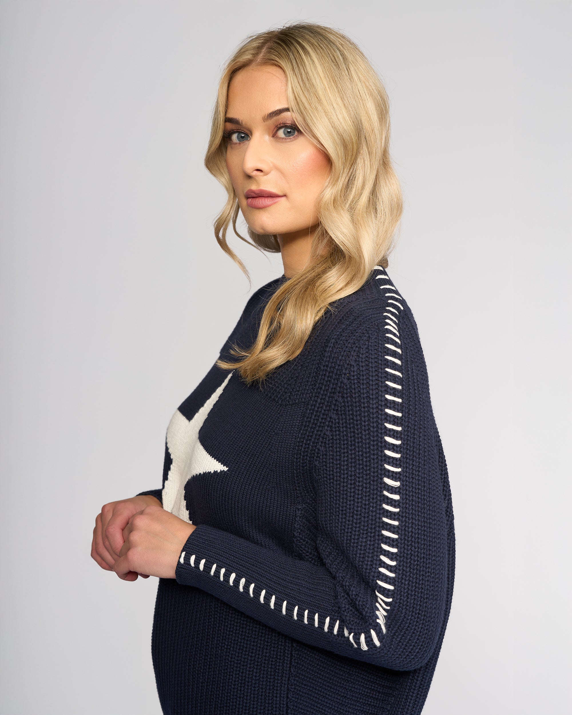 A woman with long blonde hair and light makeup wears the Vassalli Star Sweater - Ink, an oversized rib-knit navy sweater with white stitching and a large white star. She stands sideways against a plain light background, looking at the camera.