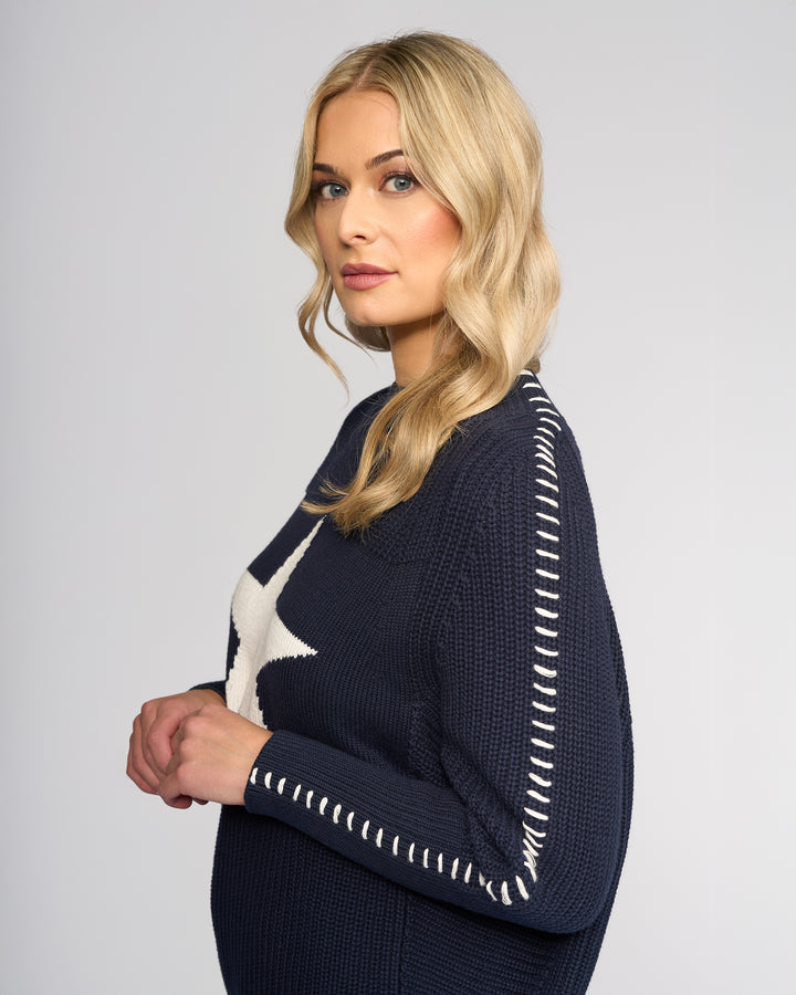 A woman with long blonde hair and light makeup wears the Vassalli Star Sweater - Ink, an oversized rib-knit navy sweater with white stitching and a large white star. She stands sideways against a plain light background, looking at the camera.