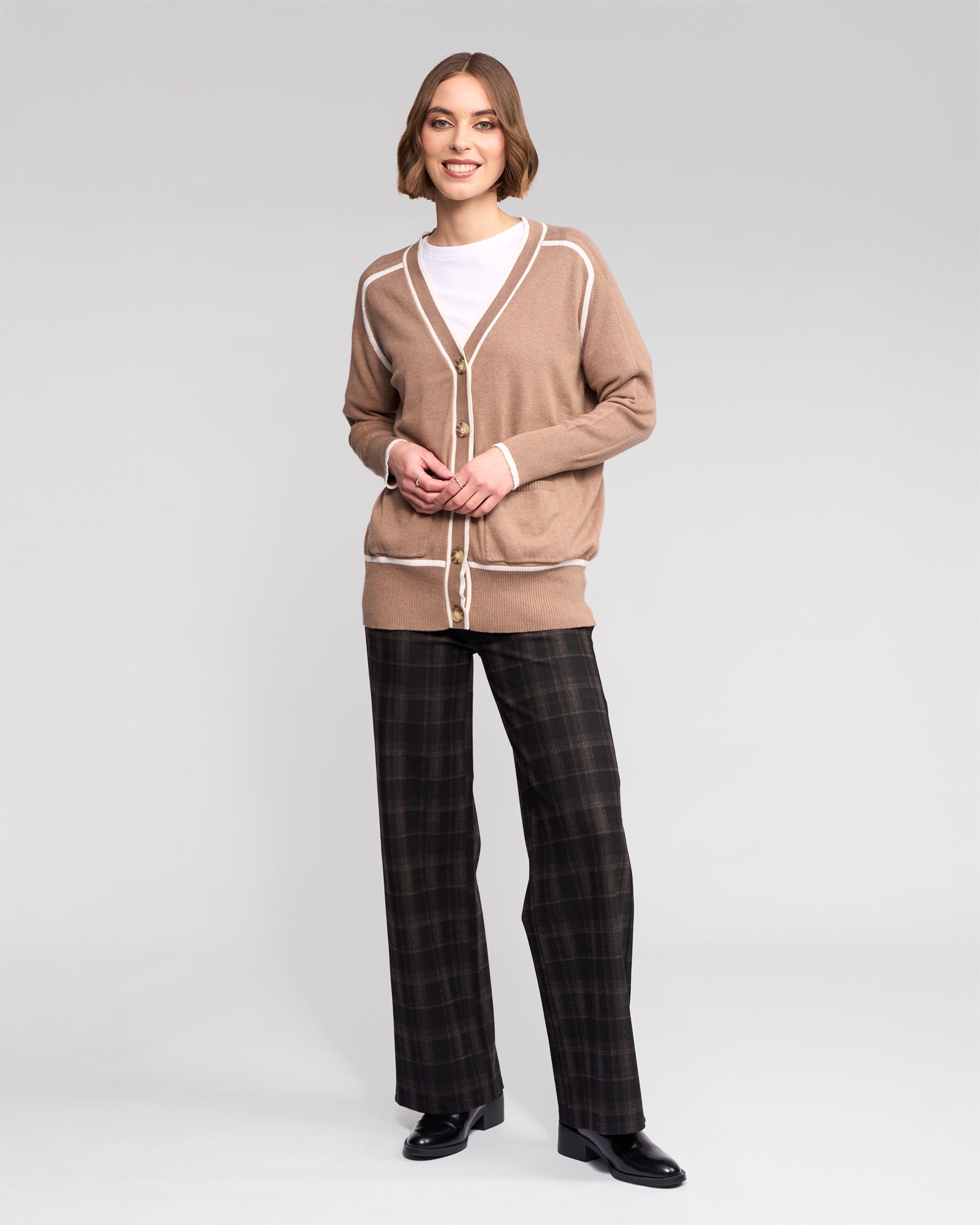 A smiling woman with short hair stands in a plain light gray studio, wearing the Vassalli Tova Cardi in Twig/Cream over a white top, paired with dark plaid trousers and black shoes. Her hands are lightly clasped.