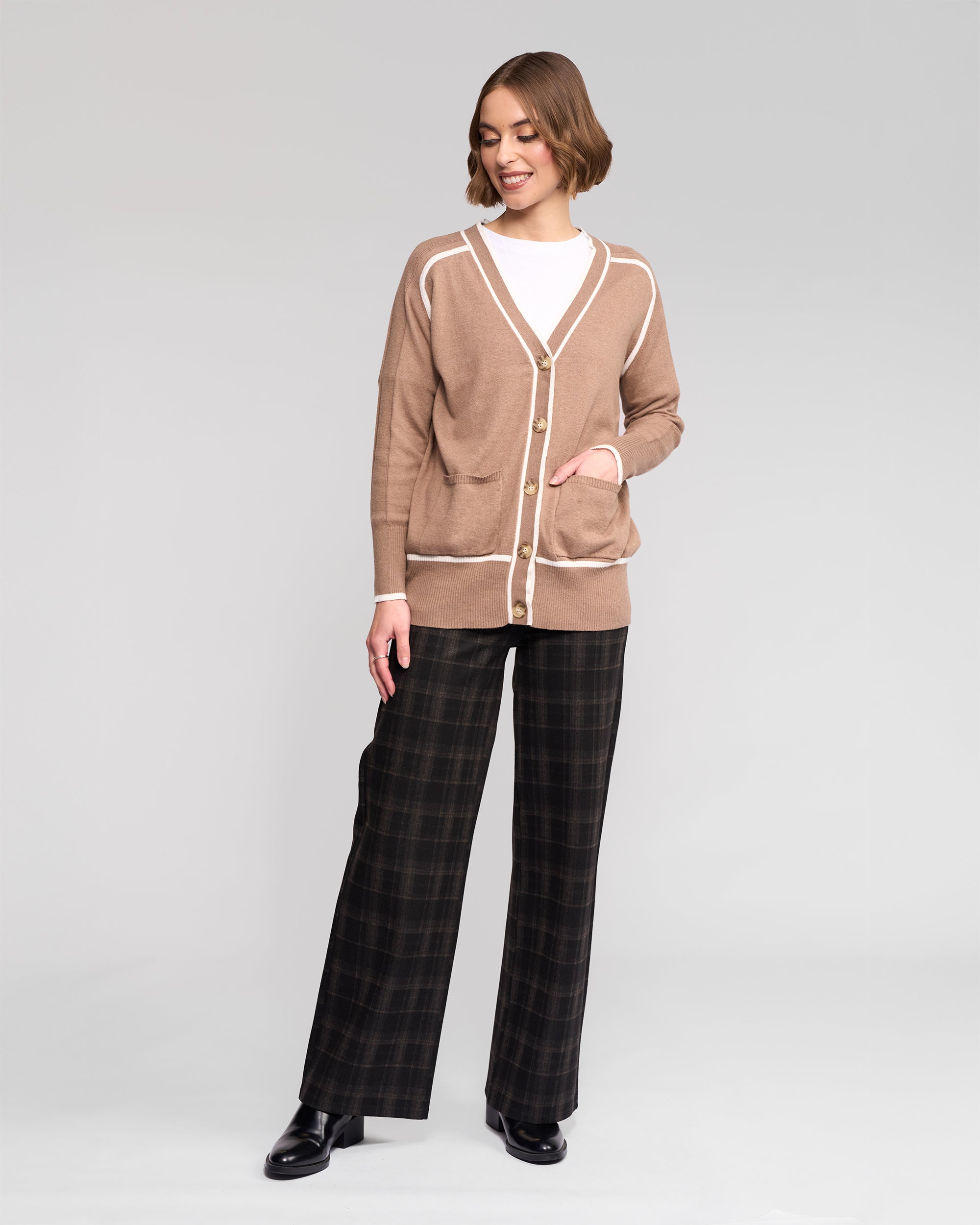 A woman stands against a plain background wearing the Vassalli Tova Cardi in Twig/Cream, a white top, wide-leg plaid pants, and black shoes. She smiles slightly and looks to the side with one hand in her cardigan pocket.