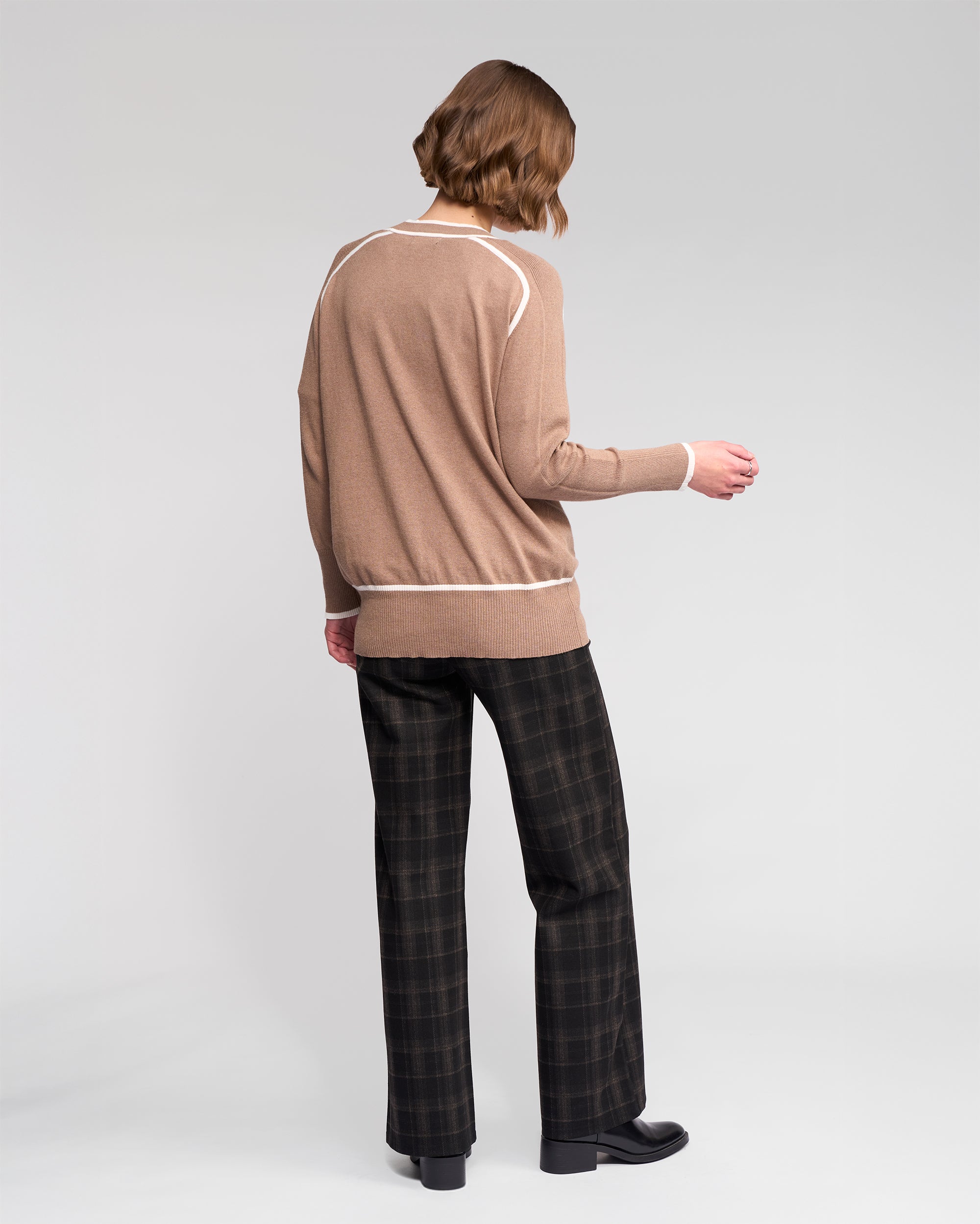 A person with short, light brown hair faces away against a plain background, wearing the Vassalli Tova Cardi in Twig/Cream, dark plaid pants, and black shoes.