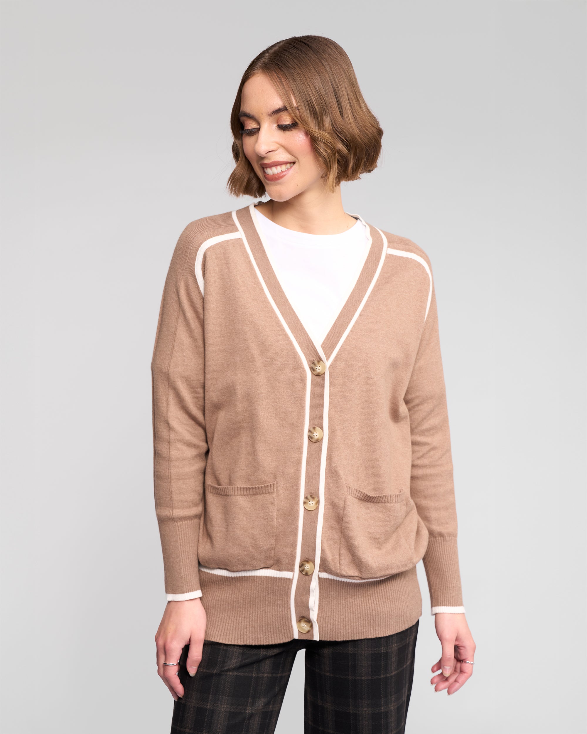 A woman with short, wavy brown hair smiles as she looks down, wearing the Vassalli Tova Cardi in Twig/Cream over a white shirt and dark plaid pants, standing against a light gray background.