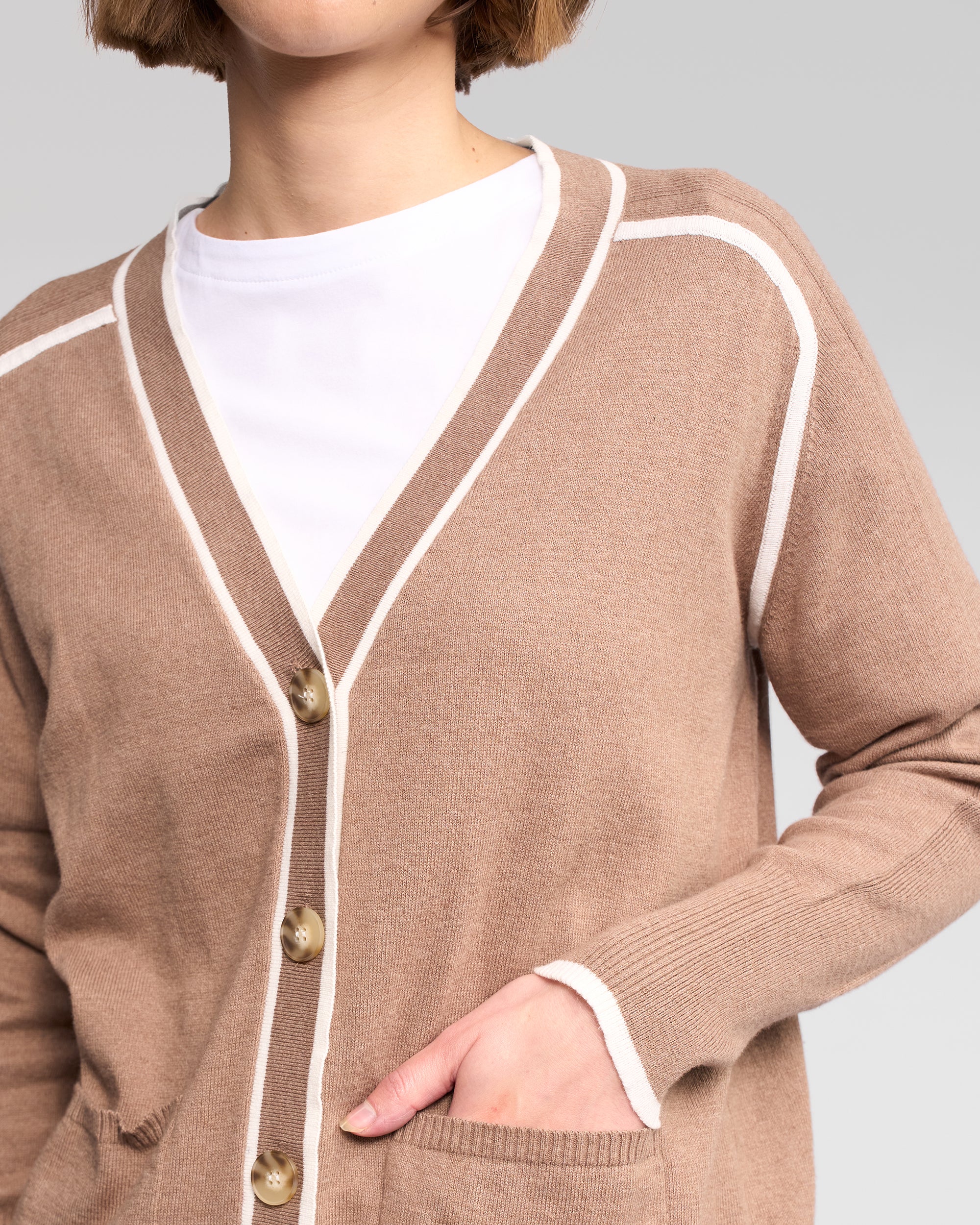 A person wears the Vassalli Tova Cardi in Twig/Cream over a white shirt, with one hand in the pocket. The image is cropped at shoulders and mid-torso.