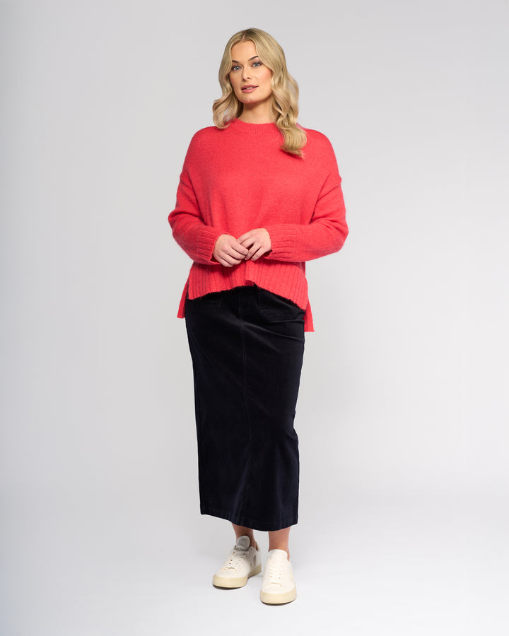 A woman with wavy blonde hair stands against a plain background, wearing the Vassalli Orbit Sweater in coral, paired with a black midi skirt and white sneakers. She has one hand in her pocket and smiles gently to the side.