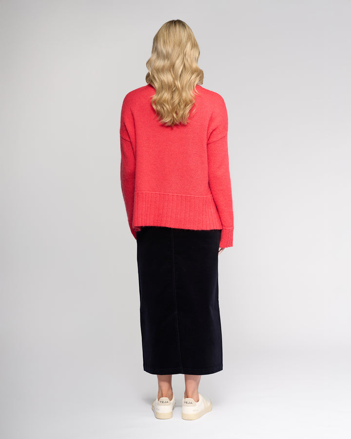A person with long wavy blonde hair stands facing away, wearing the Vassalli Orbit Sweater in Coral, paired with a black midi skirt and white sneakers against a plain light background.