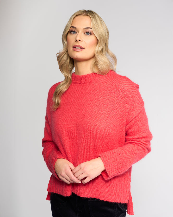 A woman with long, wavy blonde hair wears the Vassalli Orbit Sweater in Coral and black pants, standing against a plain light gray background. She looks slightly to her left with a neutral expression and her hands clasped together.