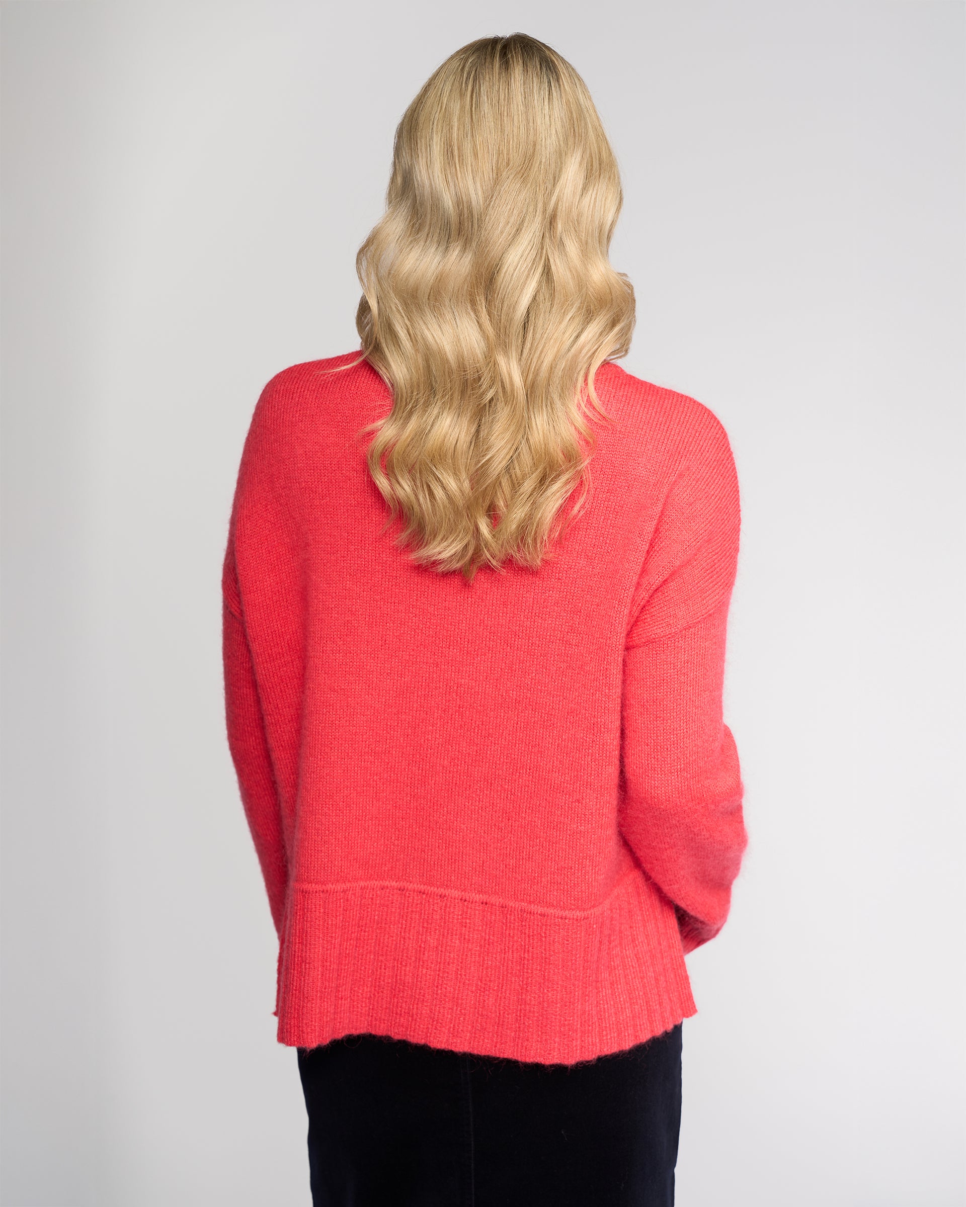 A person with long, wavy blond hair stands with their back to the camera, wearing the Vassalli Orbit Sweater in Coral and a black skirt against a plain light background.