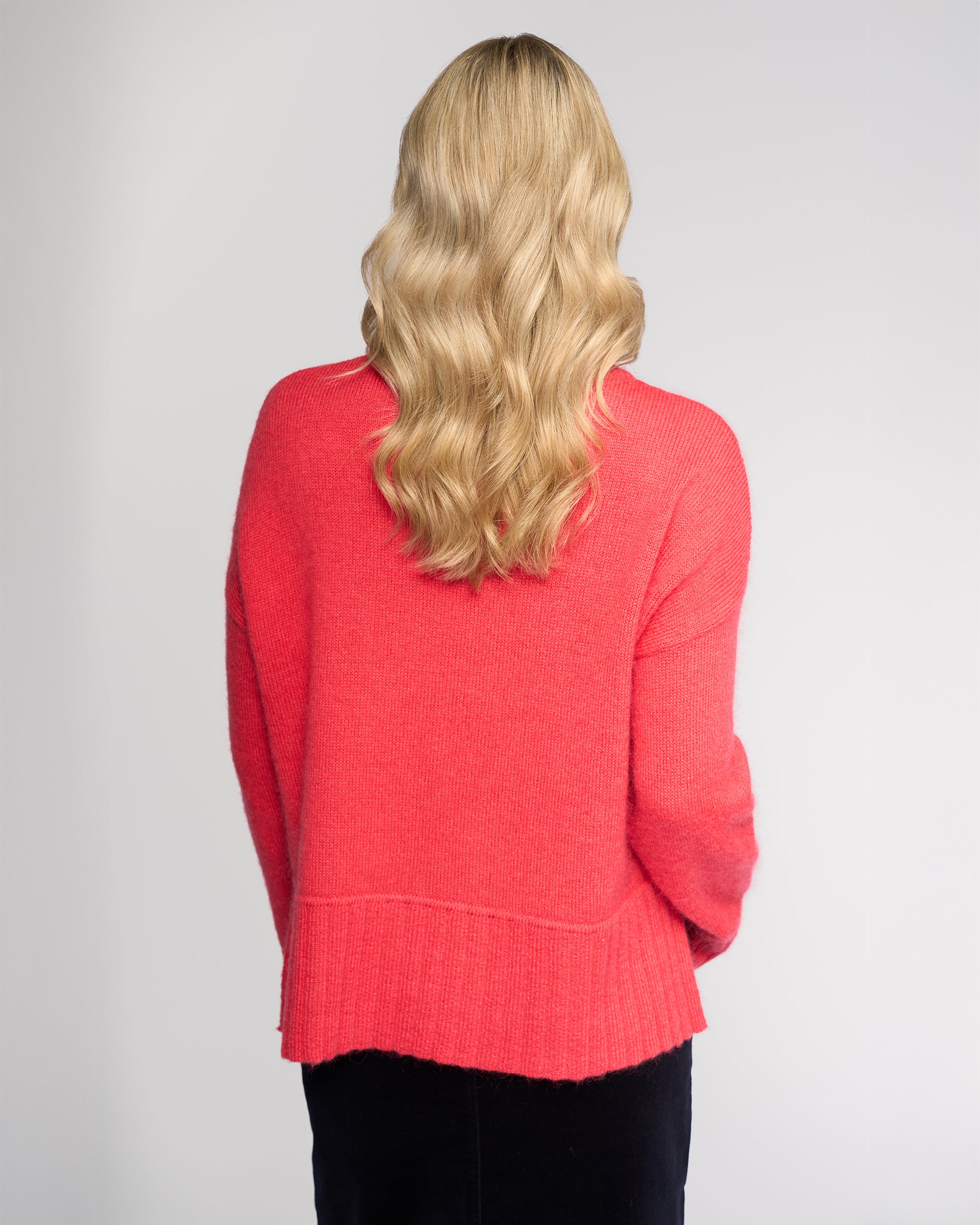 A person with long, wavy blond hair stands with their back to the camera, wearing the Vassalli Orbit Sweater in Coral and a black skirt against a plain light background.