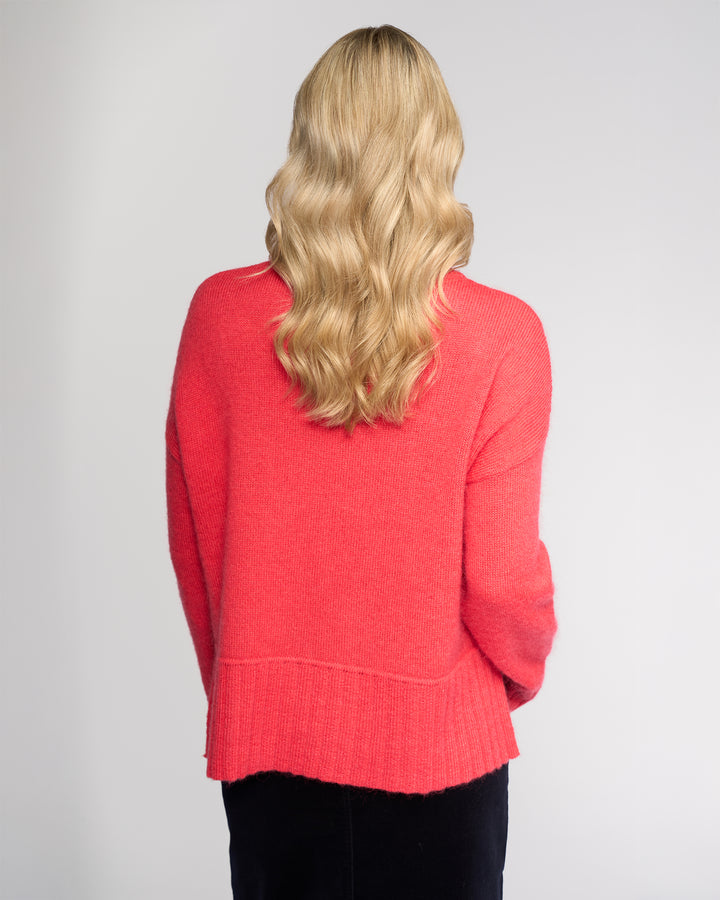 A person with long, wavy blond hair stands with their back to the camera, wearing the Vassalli Orbit Sweater in Coral and a black skirt against a plain light background.