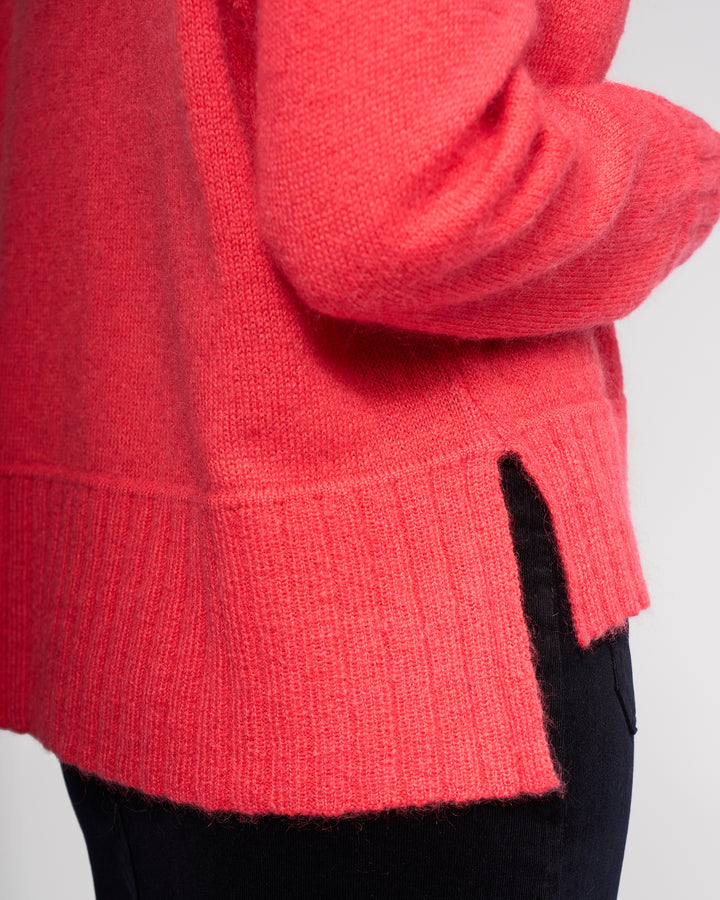 A close-up shows the torso and arm of a person wearing Vassalli’s Orbit Sweater in Coral, a cosy wool mohair blend knit with a side slit at the hem, paired with black pants.