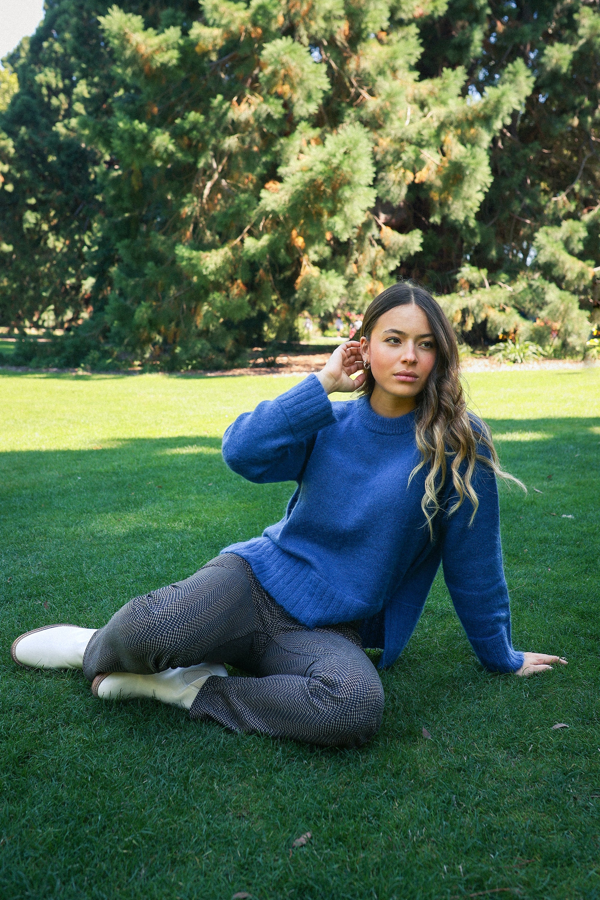 A woman with long wavy hair sits on green grass in a park, wearing the Vassalli Orbit Sweater in Denim Blue, patterned pants, and white shoes. She looks to the side with a relaxed expression as tall evergreens stand behind her.