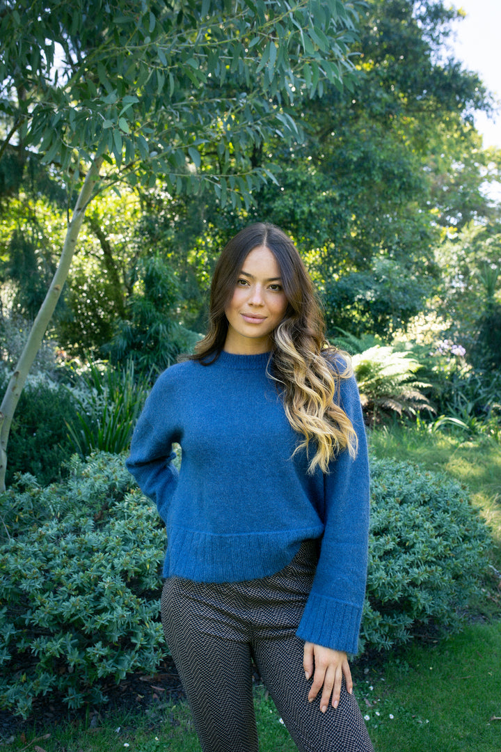 A woman with long, wavy hair stands in a lush garden, wearing the Vassalli Orbit Sweater in Denim Blue and patterned brown pants, surrounded by vibrant green plants and trees.