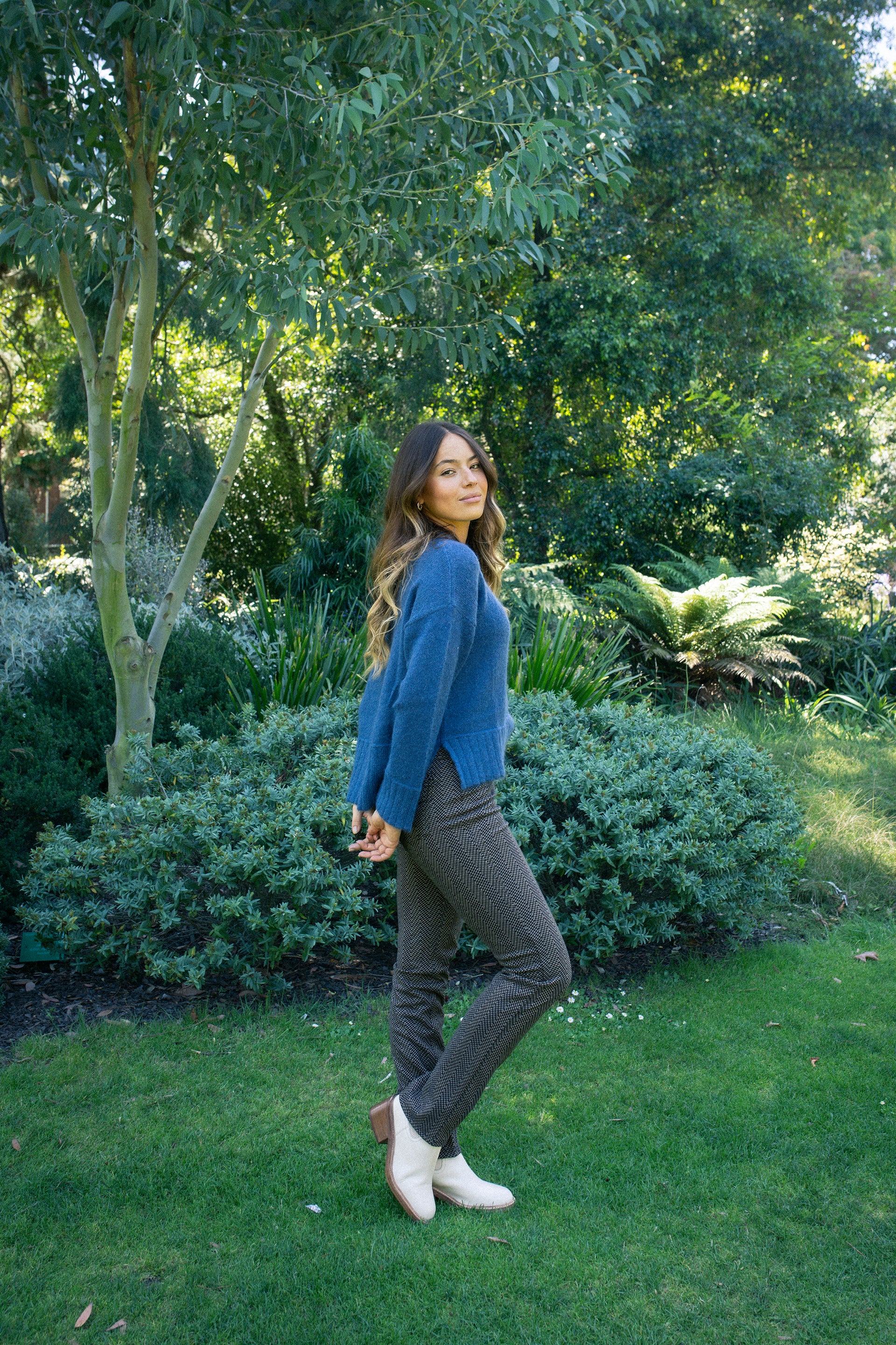 A woman with long, wavy hair stands on green grass in a garden, wearing Vassalli's Bay Pull On - Arc slim pants and a blue sweater with white ankle boots. She smiles and looks to the side, surrounded by lush greenery and trees.