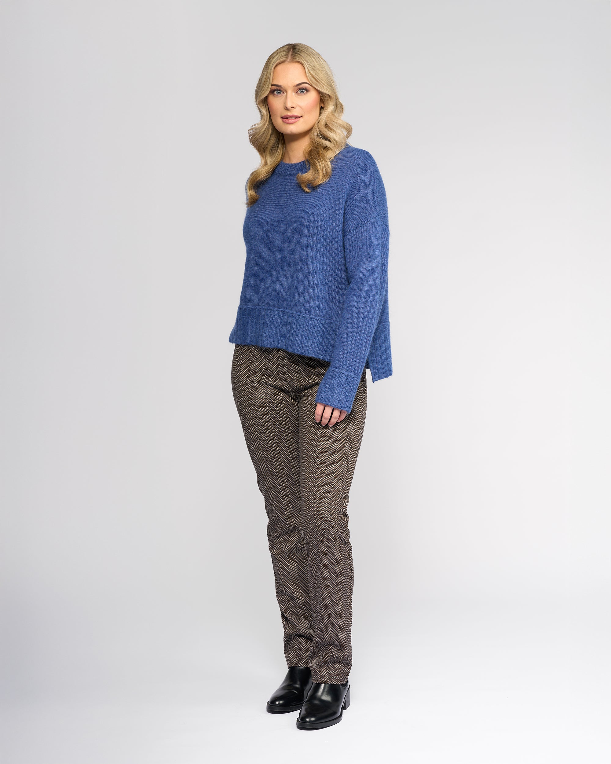 A woman with blonde hair wears the Vassalli Orbit Sweater in Denim Blue, featuring a relaxed fit and wool-mohair blend. She pairs it with patterned brown pants and black shoes, standing against a plain white background facing forward.