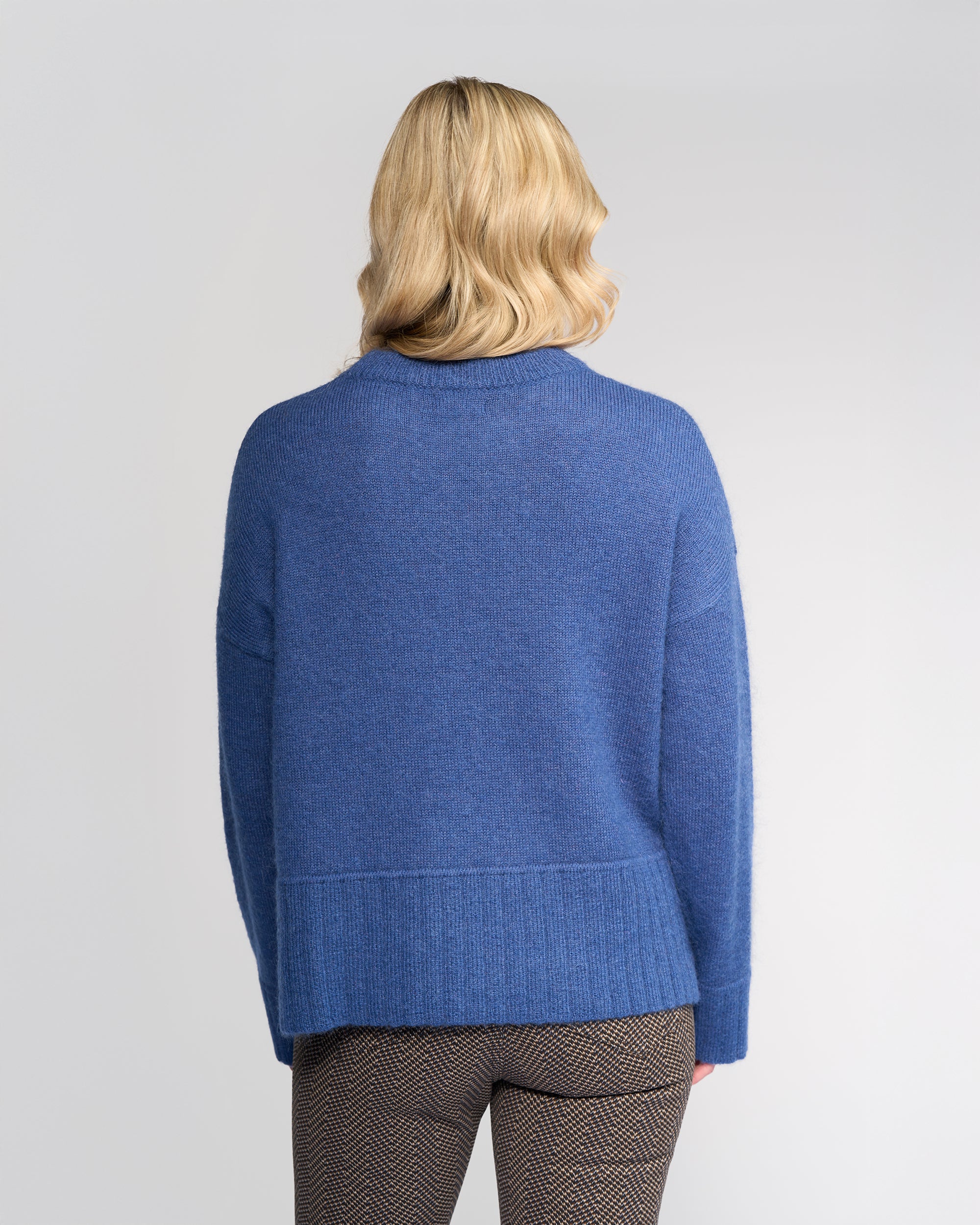A person with wavy blonde hair is seen from behind, wearing the Vassalli Orbit Sweater in Denim Blue and brown patterned pants, standing against a plain light background.