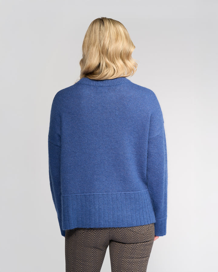 A person with wavy blonde hair is seen from behind, wearing the Vassalli Orbit Sweater in Denim Blue and brown patterned pants, standing against a plain light background.