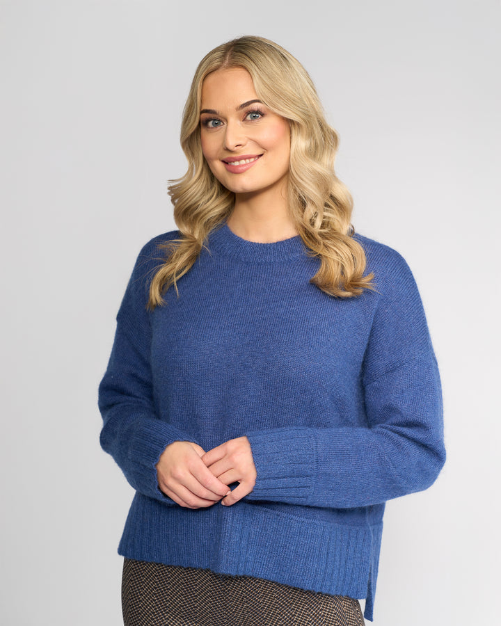 A woman with long blonde hair smiles warmly, standing against a light gray background in the Vassalli Orbit Sweater – Denim Blue, her hands gently clasped in front.