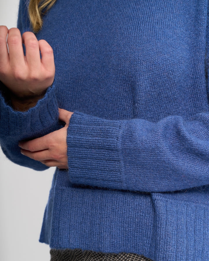 Close-up of a person wearing the Vassalli Orbit Sweater in Denim Blue, relaxed-fit knit, gently holding their right sleeve near the wrist. Only the torso and arms are shown.