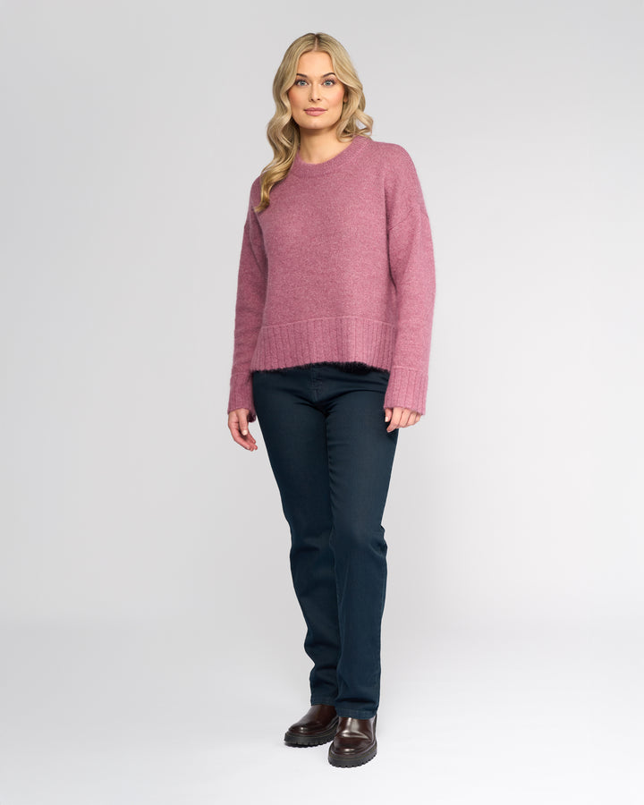 A woman with wavy blonde hair wears the Vassalli Orbit Sweater in Port, a relaxed-fit knit, styled with dark blue jeans and brown shoes. She stands against a plain light gray background with a neutral expression.