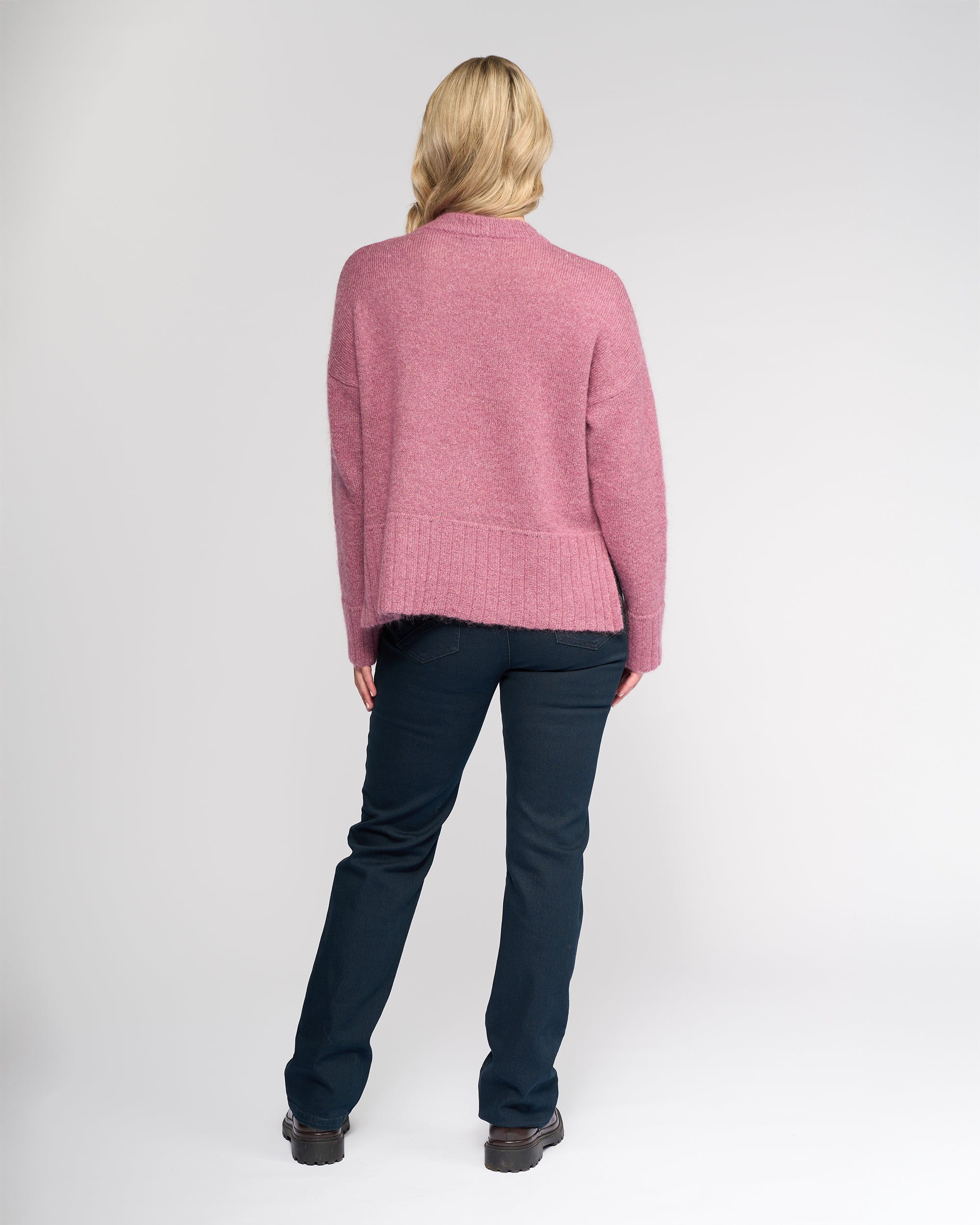 A person with blonde hair faces away, wearing the Vassalli Orbit Sweater in Port, paired with dark blue jeans and black shoes against a plain light background.