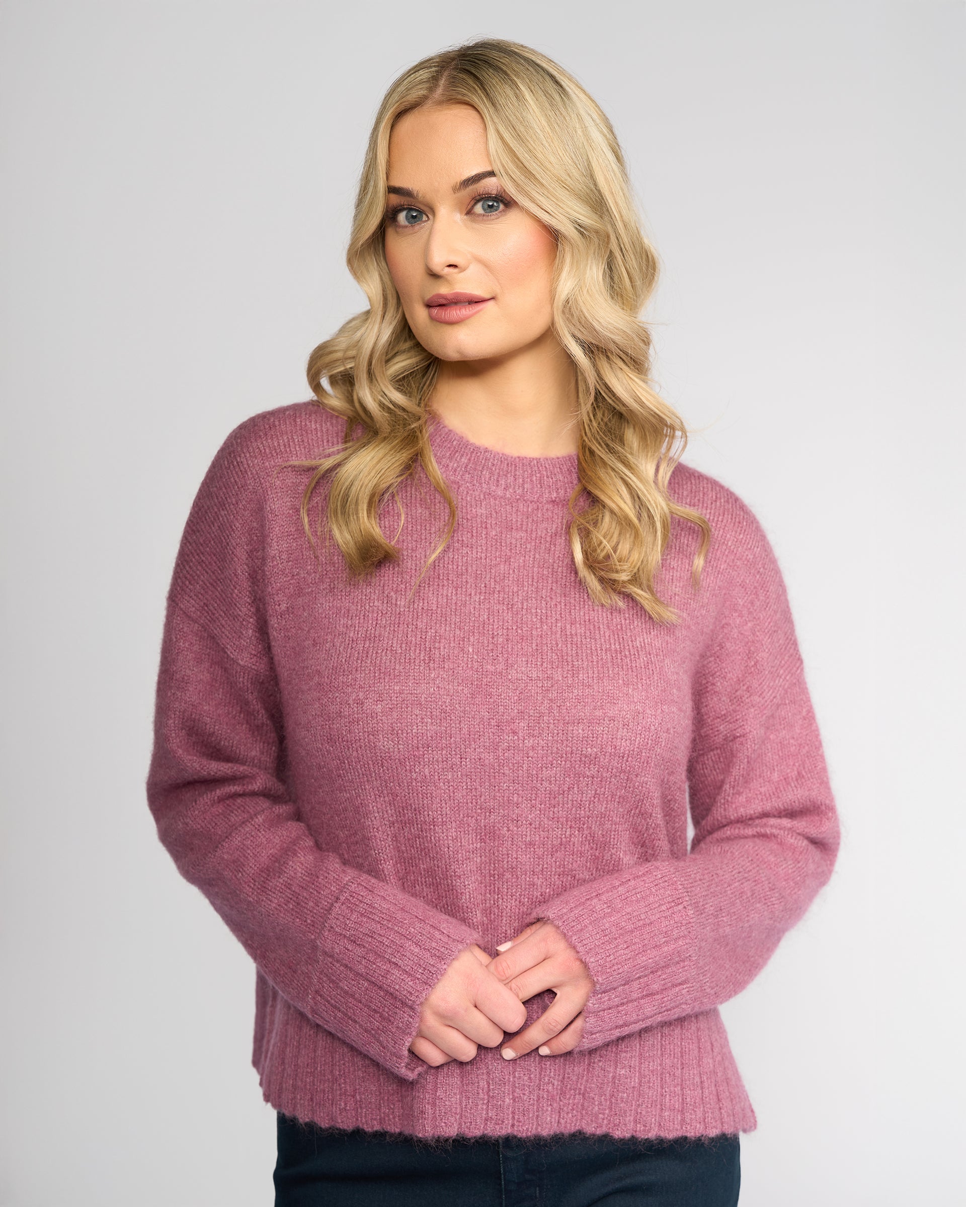 A woman with wavy blonde hair wears the Vassalli Orbit Sweater in Port, a relaxed-fit knit, standing against a plain light gray background with her hands clasped and a neutral expression.