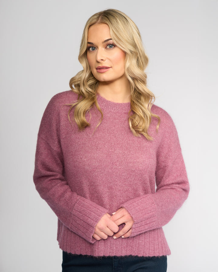 A woman with wavy blonde hair wears the Vassalli Orbit Sweater in Port, a relaxed-fit knit, standing against a plain light gray background with her hands clasped and a neutral expression.
