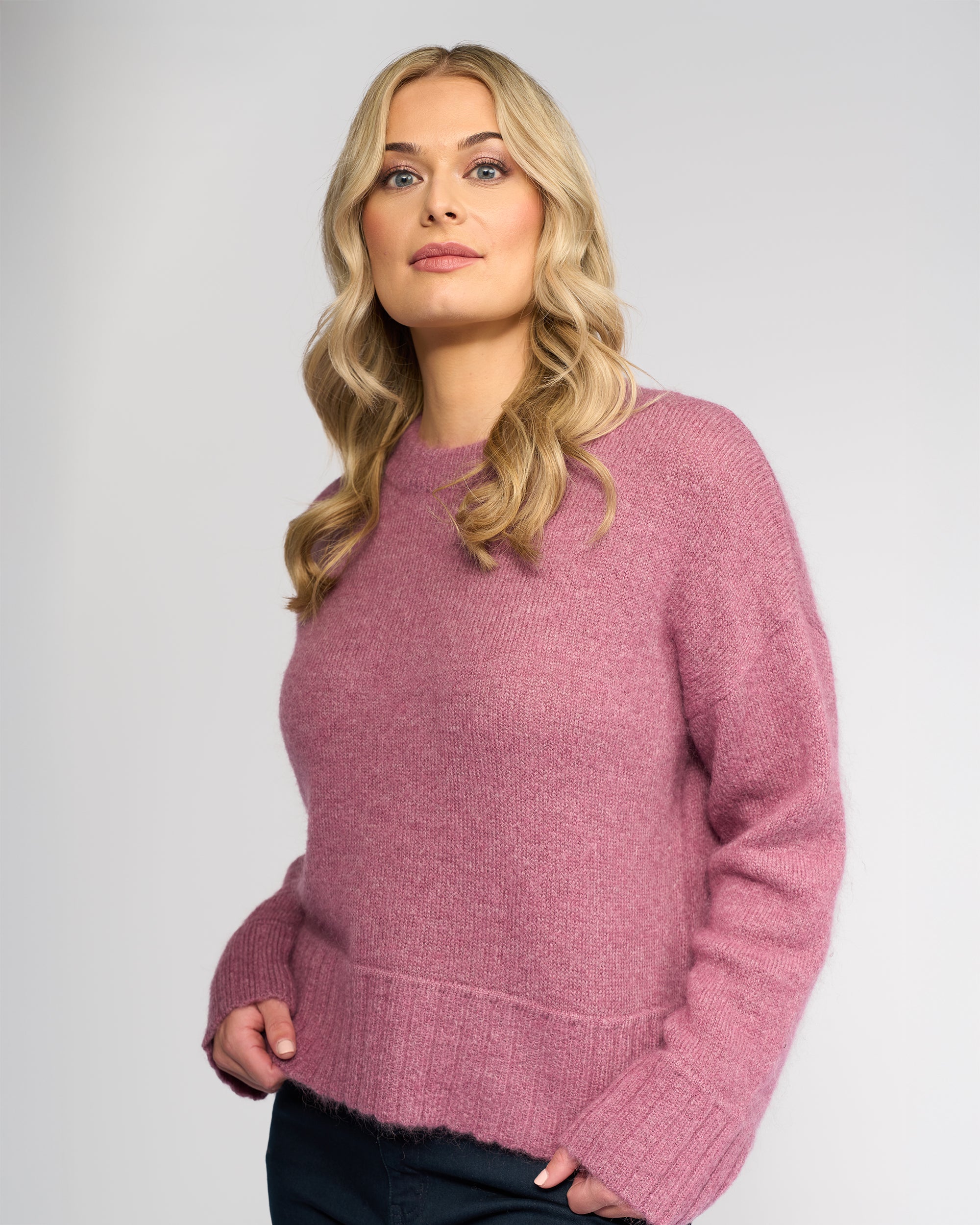A woman with long blonde hair wears the cosy, relaxed-fit Orbit Sweater in Port by Vassalli, standing against a plain light gray background and looking confidently at the camera with a slight smile.