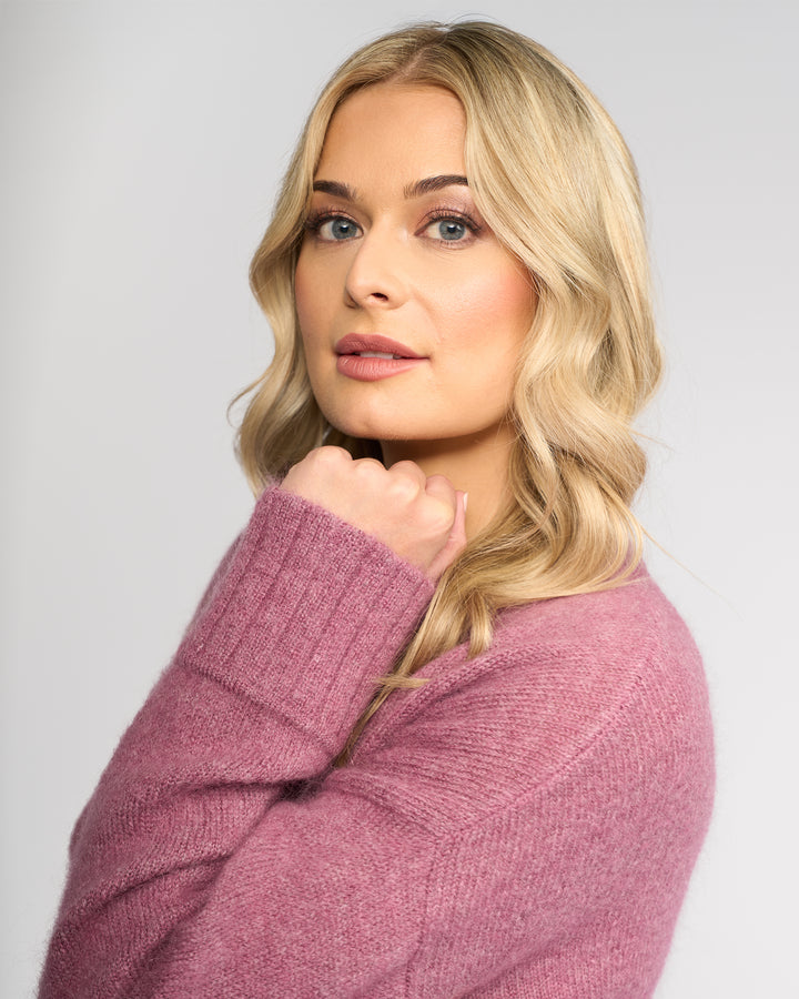 A woman with long, wavy blonde hair models the Vassalli Orbit Sweater in Port, exuding confidence as she poses with her hand near her chin against a light gray background.