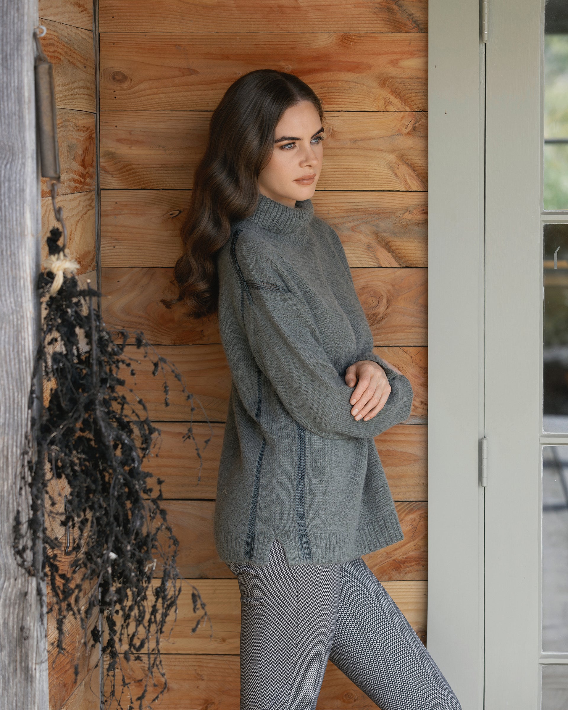 A woman with long wavy brown hair, arms crossed, wears the Vassalli Mellow Sweater - Elm and patterned pants, leaning against a wooden wall by a door. A bundle of dried black plants hangs on the left.