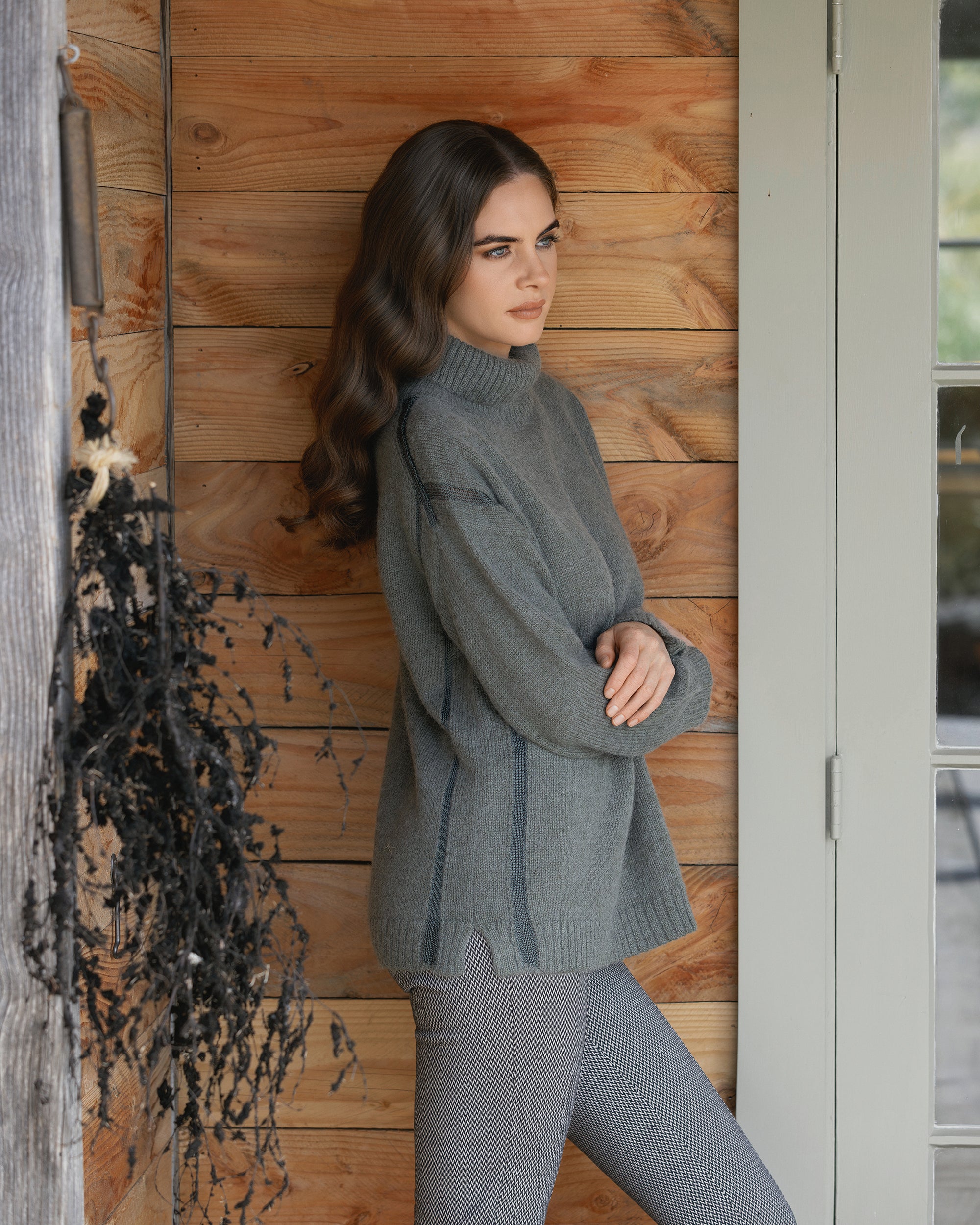 A woman with long wavy brown hair, arms crossed, wears the Vassalli Mellow Sweater - Elm and patterned pants, leaning against a wooden wall by a door. A bundle of dried black plants hangs on the left.