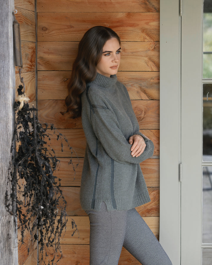 A woman with long wavy brown hair, arms crossed, wears the Vassalli Mellow Sweater - Elm and patterned pants, leaning against a wooden wall by a door. A bundle of dried black plants hangs on the left.