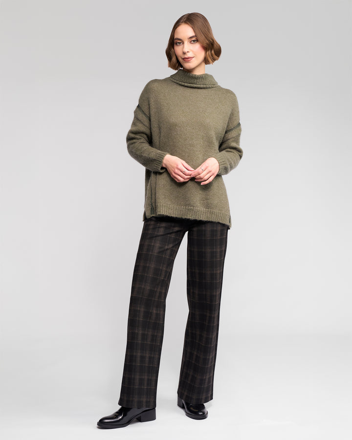 A person stands against a plain light gray background wearing the Vassalli Mellow Sweater in Elm, paired with dark plaid trousers and black shoes, arms relaxed and a slight smile.
