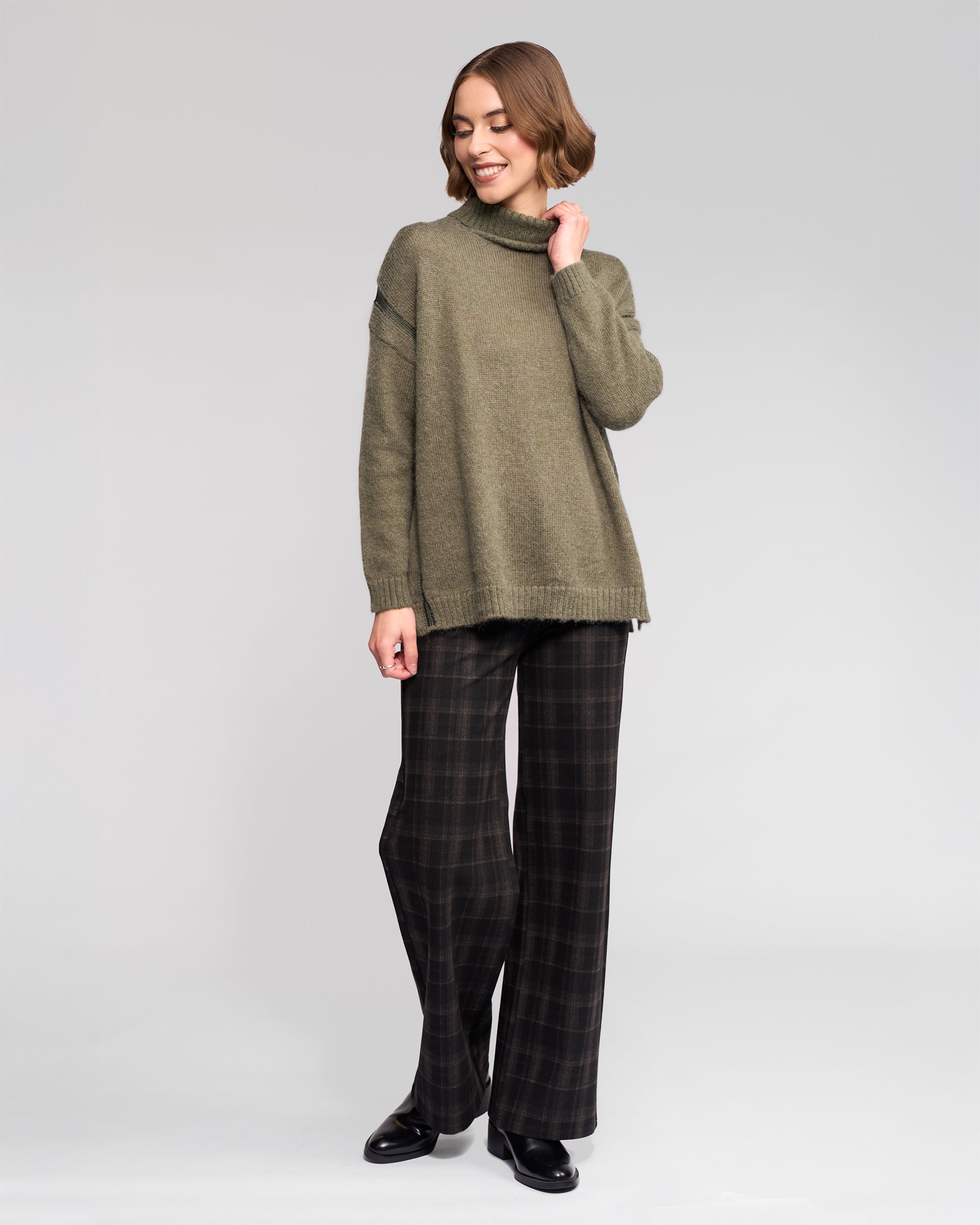 A woman smiles in the Vassalli Mellow Sweater - Elm, an oversized olive-green mohair wool turtleneck, paired with wide-legged dark plaid pants and black shoes, standing against a plain light gray background.