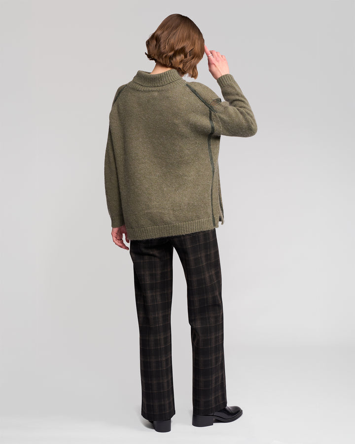 A person faces away from the camera wearing the Vassalli Mellow Sweater in Elm, paired with dark plaid pants and black shoes, one hand raised near their head against a plain light background.