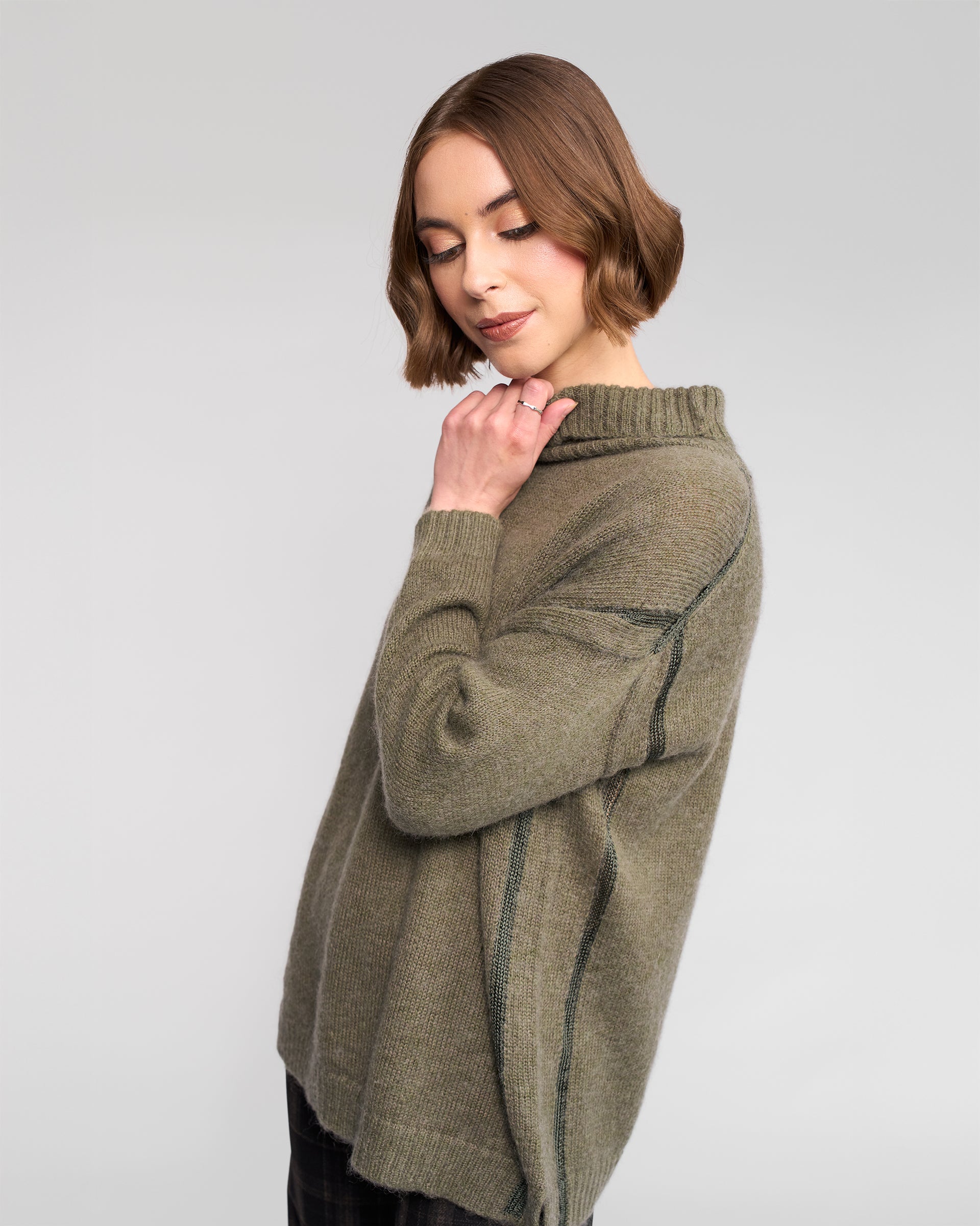 A woman with short brown hair models the Vassalli Mellow Sweater in Elm, standing in profile against a light gray background, gently touching her chin and looking down with a soft expression.