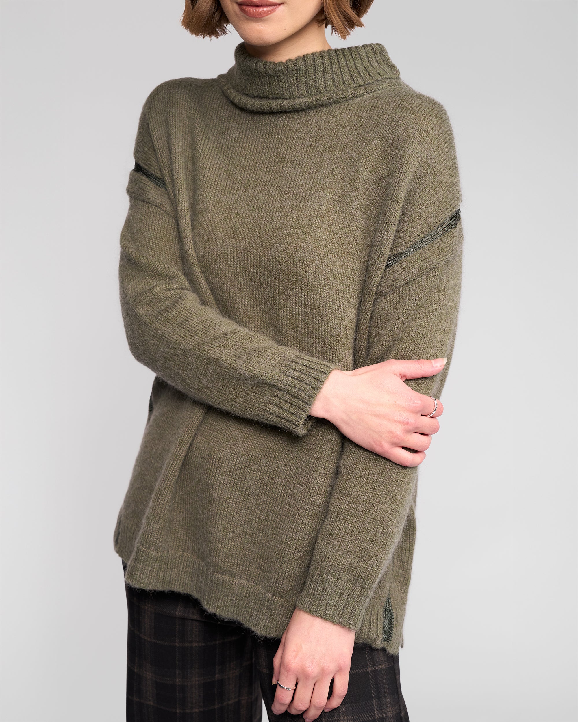 A woman stands with arms crossed, wearing the Vassalli Mellow Sweater - Elm, an olive green mohair wool turtleneck with long sleeves, paired with black plaid pants. Her face is partially out of the frame.