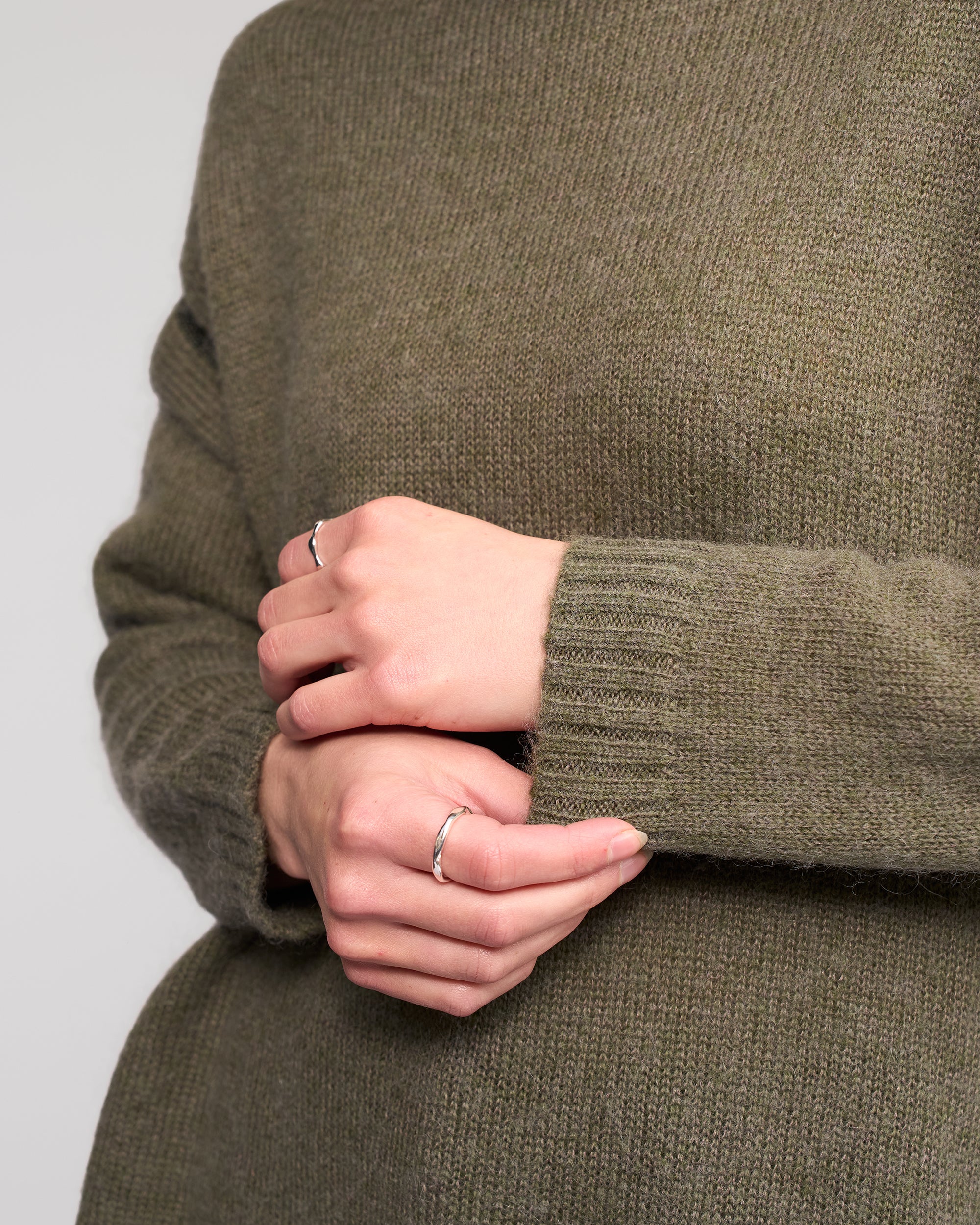 A person wearing the Vassalli Mellow Sweater in Elm, shown from the shoulders down, gently holds one hand with the other; both hands feature simple silver rings.