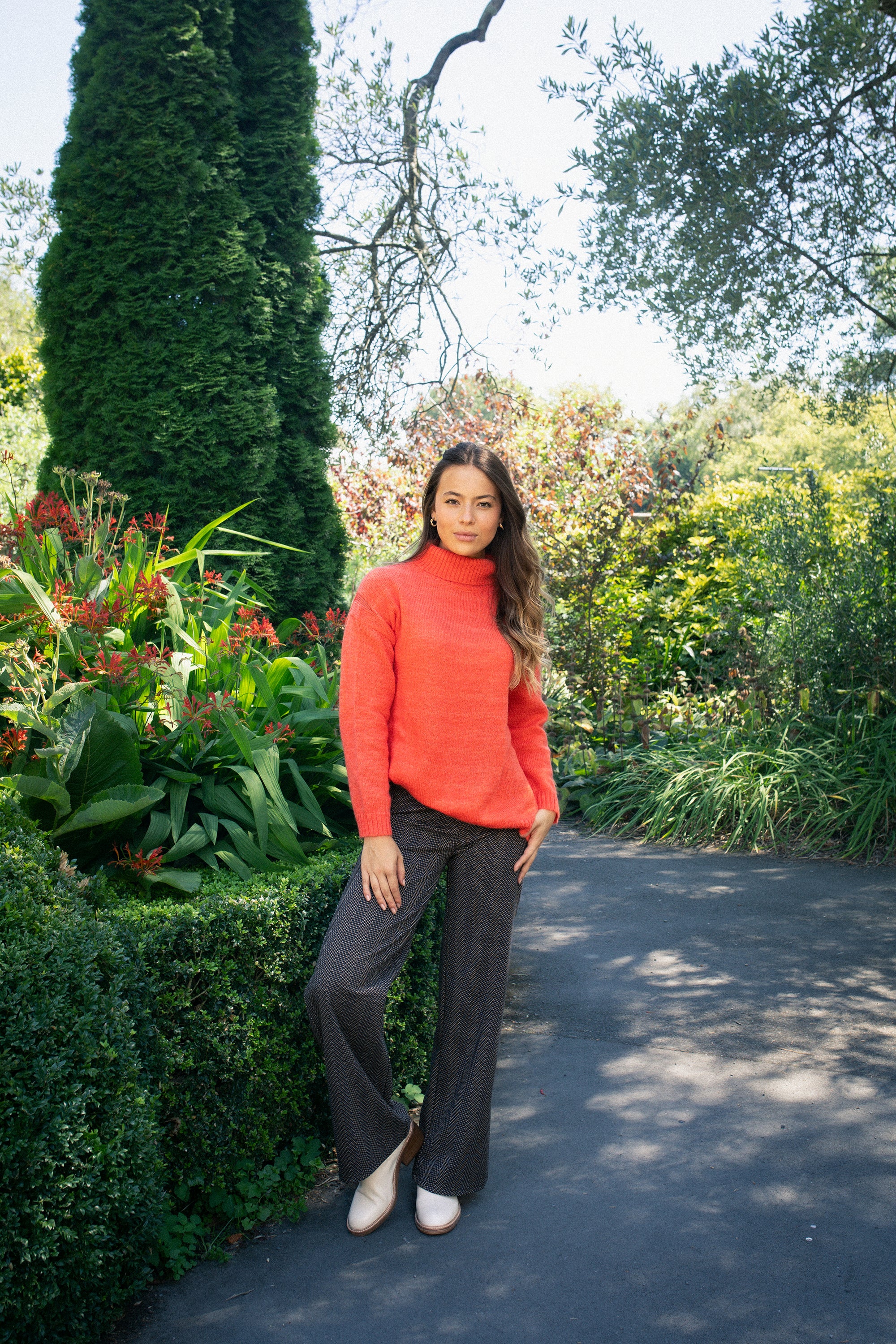 A woman stands on a sunny garden path in lush greenery, wearing Vassalli's Loft Pull On - Arc dark wide-leg pants with an elastic waistband, a red stretch knit turtleneck sweater, and white shoes.