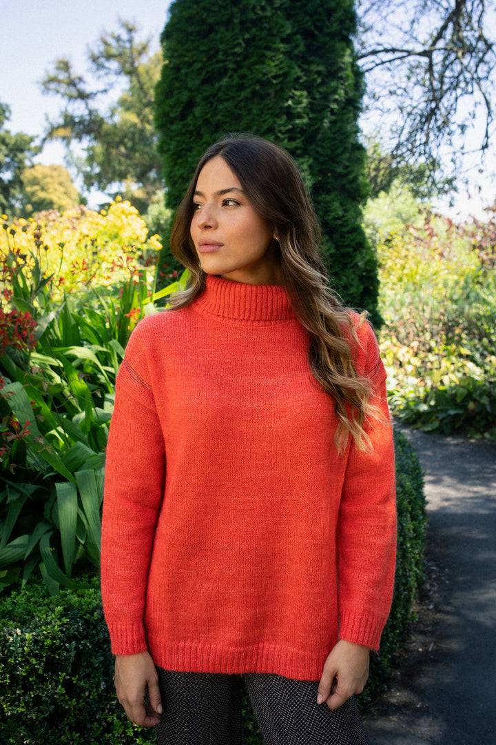 A woman with long wavy hair stands outdoors in a lush garden, wearing the Vassalli Mellow Sweater - Lava, a bright orange mohair wool piece with shiny thread detailing, and looks off to the side amid green plants and flowers.