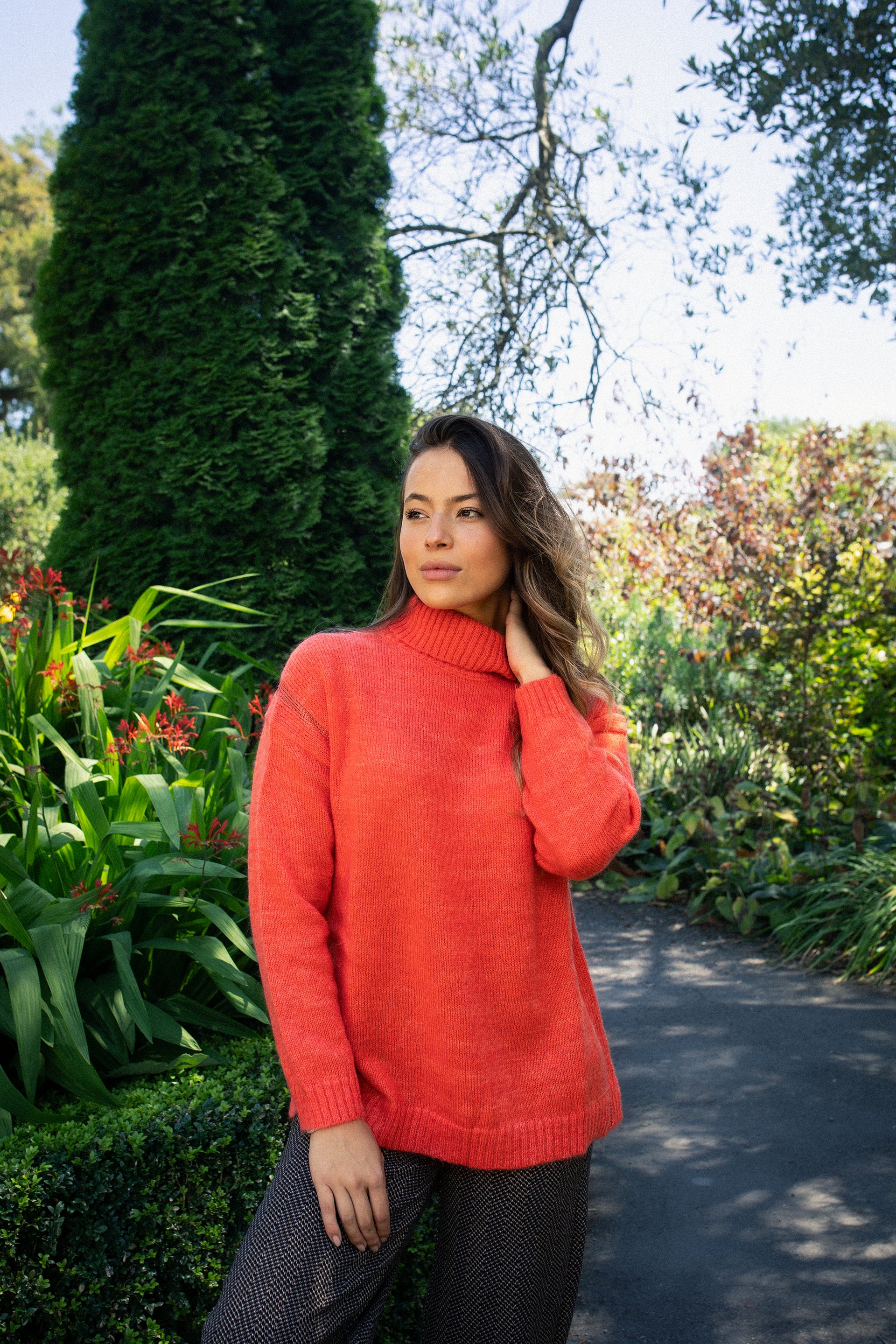 A woman wearing the Vassalli Mellow Sweater in Lava stands outdoors among lush green plants and flowers, touching her hair with a thoughtful look.