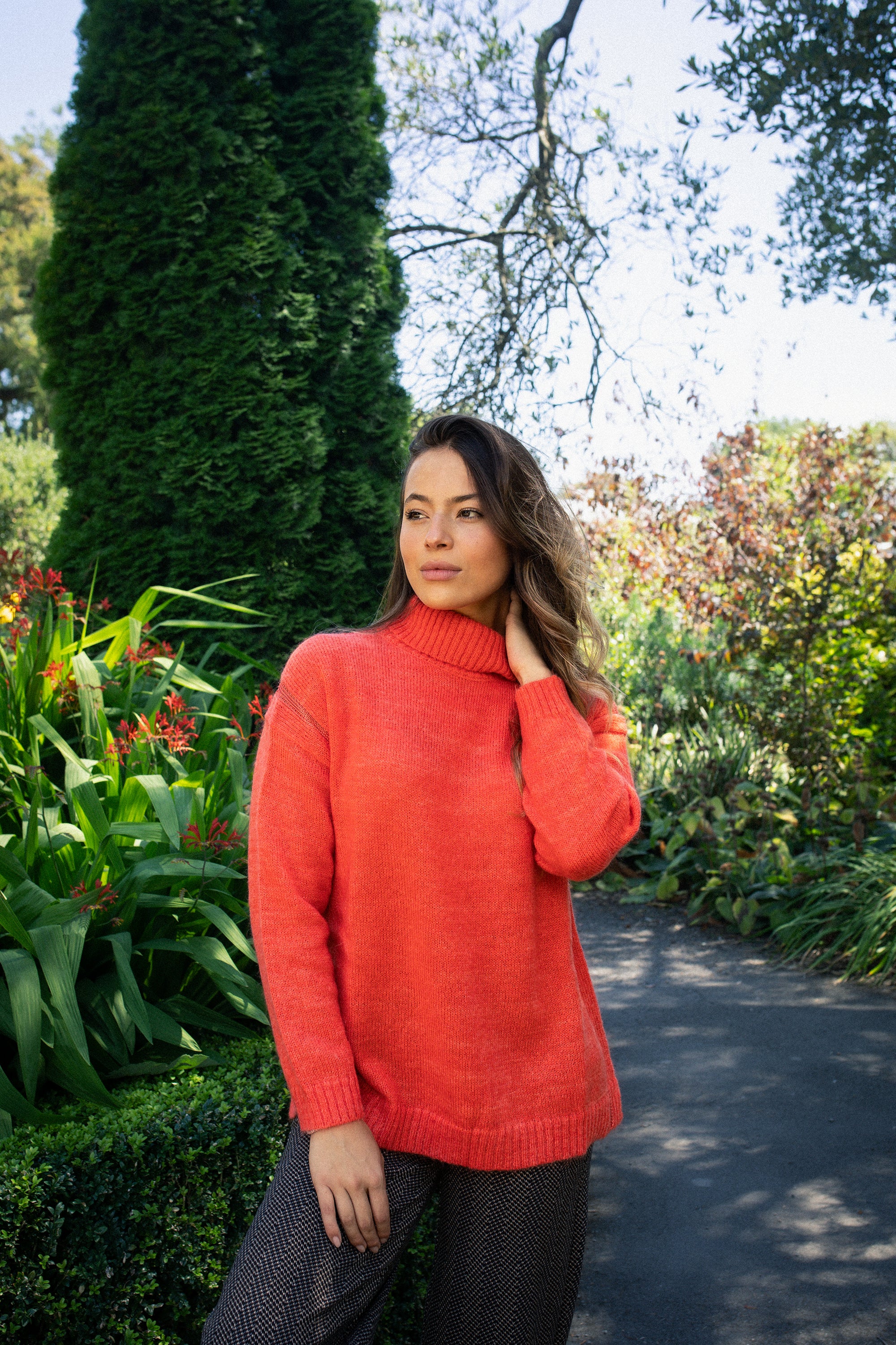 A woman wearing the Vassalli Mellow Sweater in Lava stands outdoors among lush green plants and flowers, touching her hair with a thoughtful look.