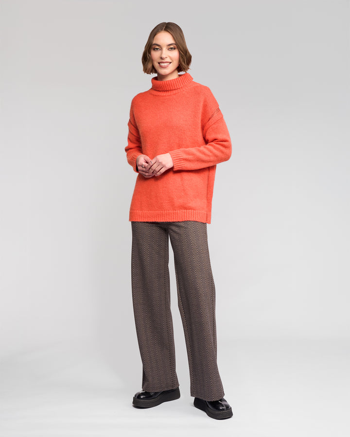 A woman smiles in the Vassalli Mellow Sweater - Lava, paired with brown patterned wide-leg pants and black shoes, set against a light gray background.