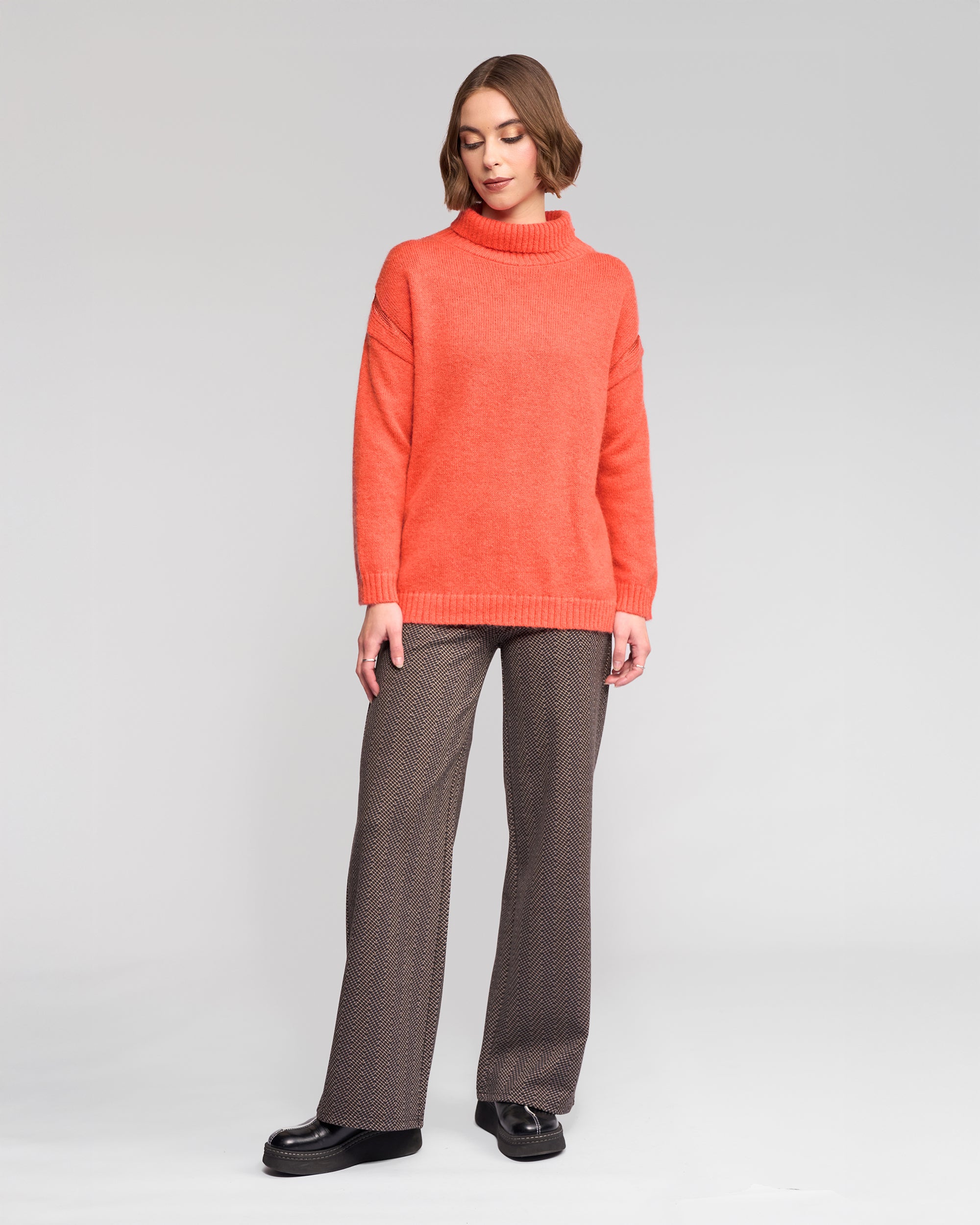 A woman with short brown hair poses against a plain background wearing Vassalli’s Mellow Sweater in Lava, styled with patterned brown trousers and black platform shoes.