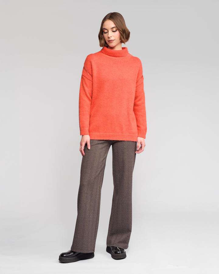 A woman with short brown hair poses against a plain background wearing Vassalli’s Mellow Sweater in Lava, styled with patterned brown trousers and black platform shoes.