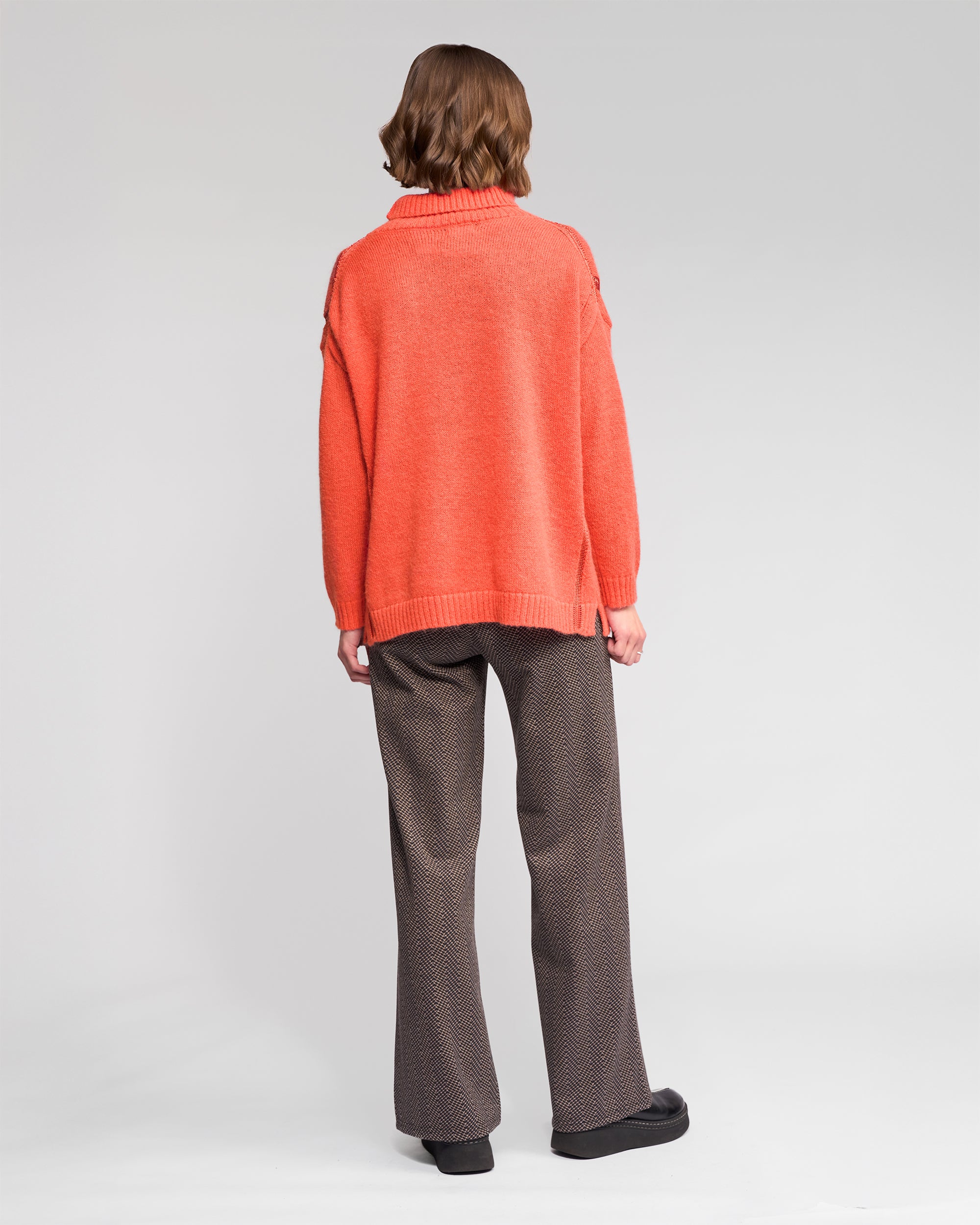 A person with short, wavy brown hair stands facing away, wearing the Vassalli Mellow Sweater - Lava, along with brown patterned pants and black shoes against a plain, light background.