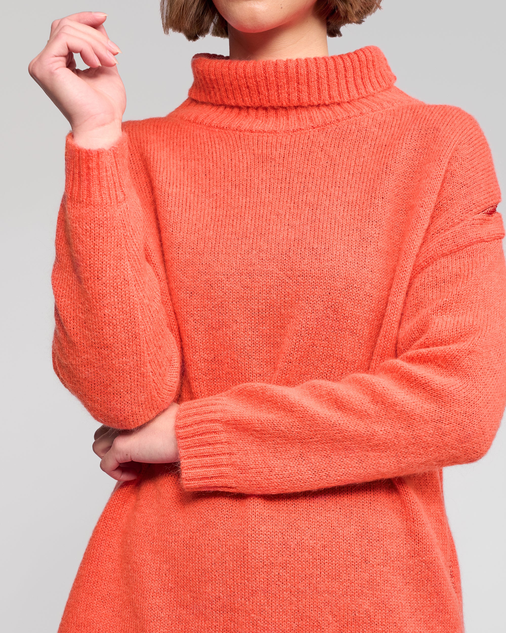 A person with short hair wears the Vassalli Mellow Sweater - Lava, standing with one arm crossed and the other hand raised near their face; their face is partially out of frame.