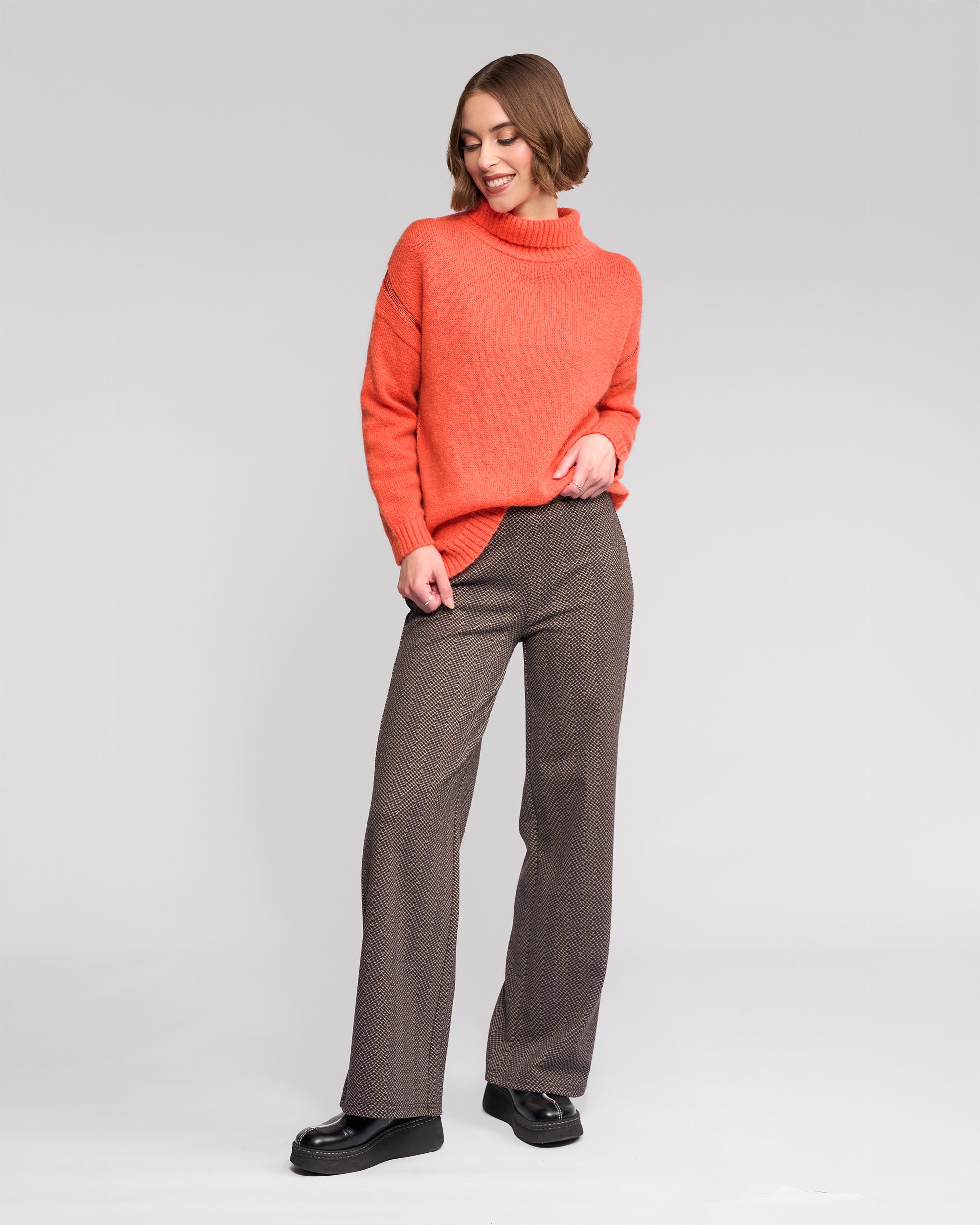 A person stands against a plain background, wearing the Vassalli Mellow Sweater - Lava, paired with patterned brown wide-leg trousers and black shoes, looking down and smiling with one hand in their pocket.
